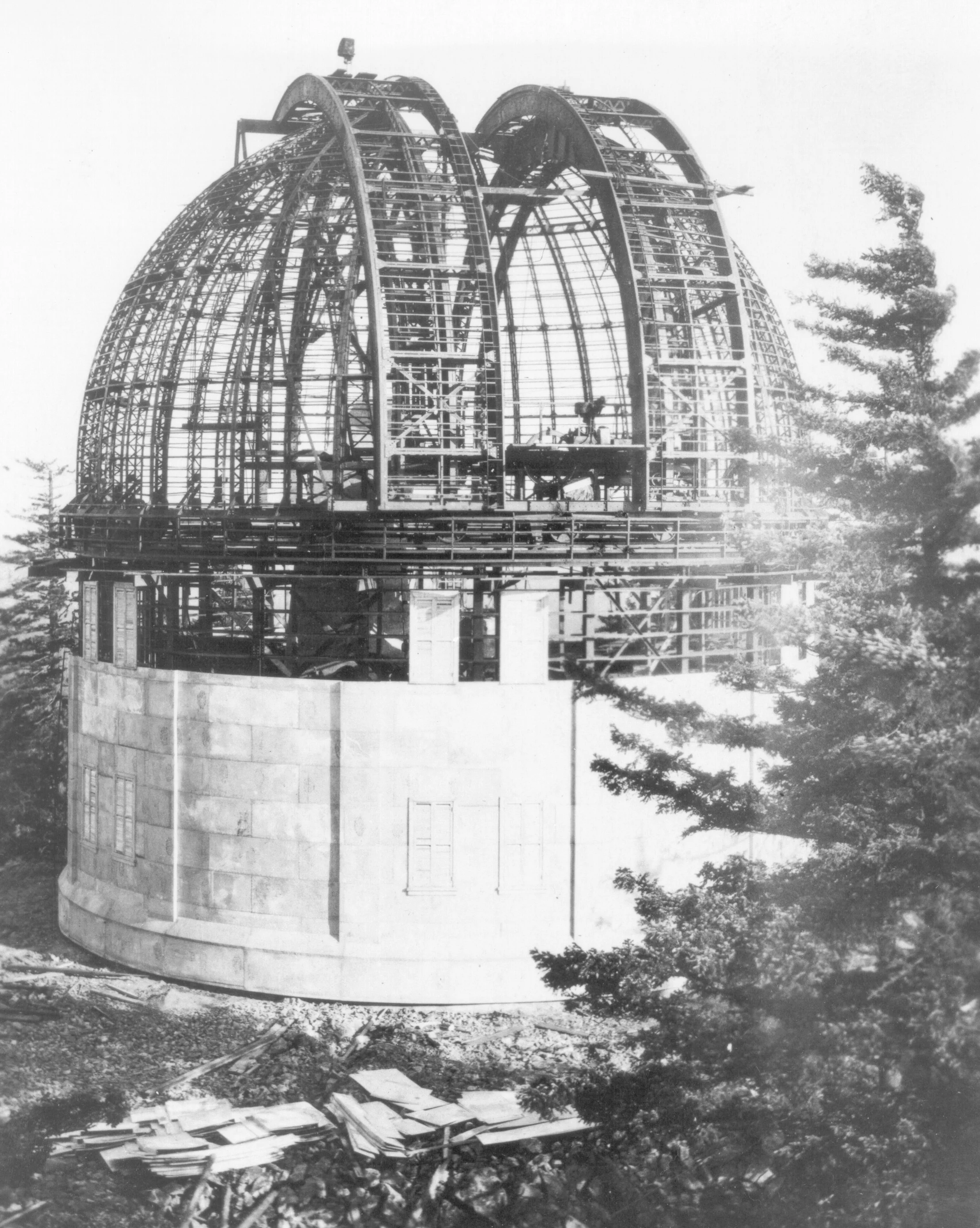Historical photo of an observatory under construction with a dome framework, surrounded by trees.