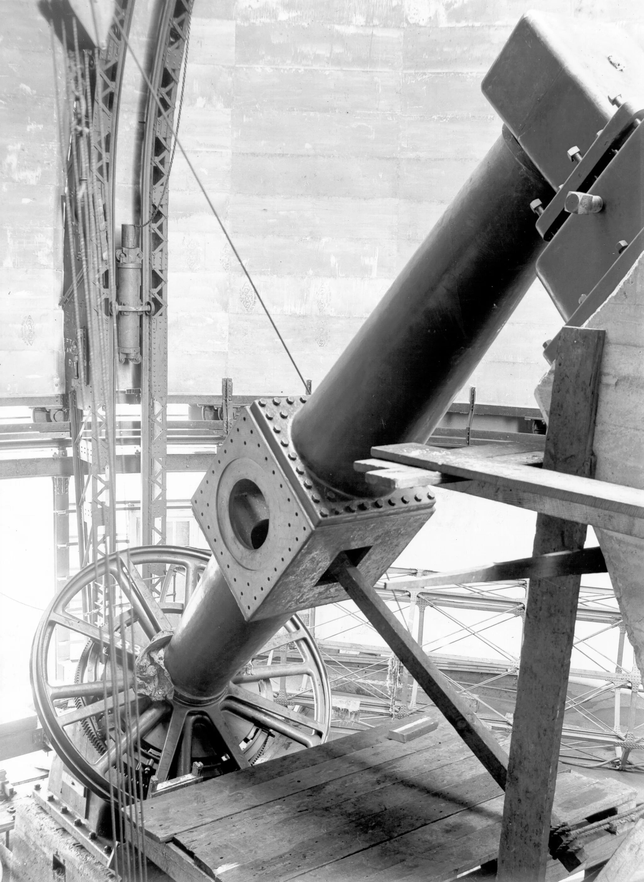 Black and white photo of a large industrial pipe with flanged ends, supported by wooden planks on a construction site. Surrounding scaffolding and metal framework are visible.