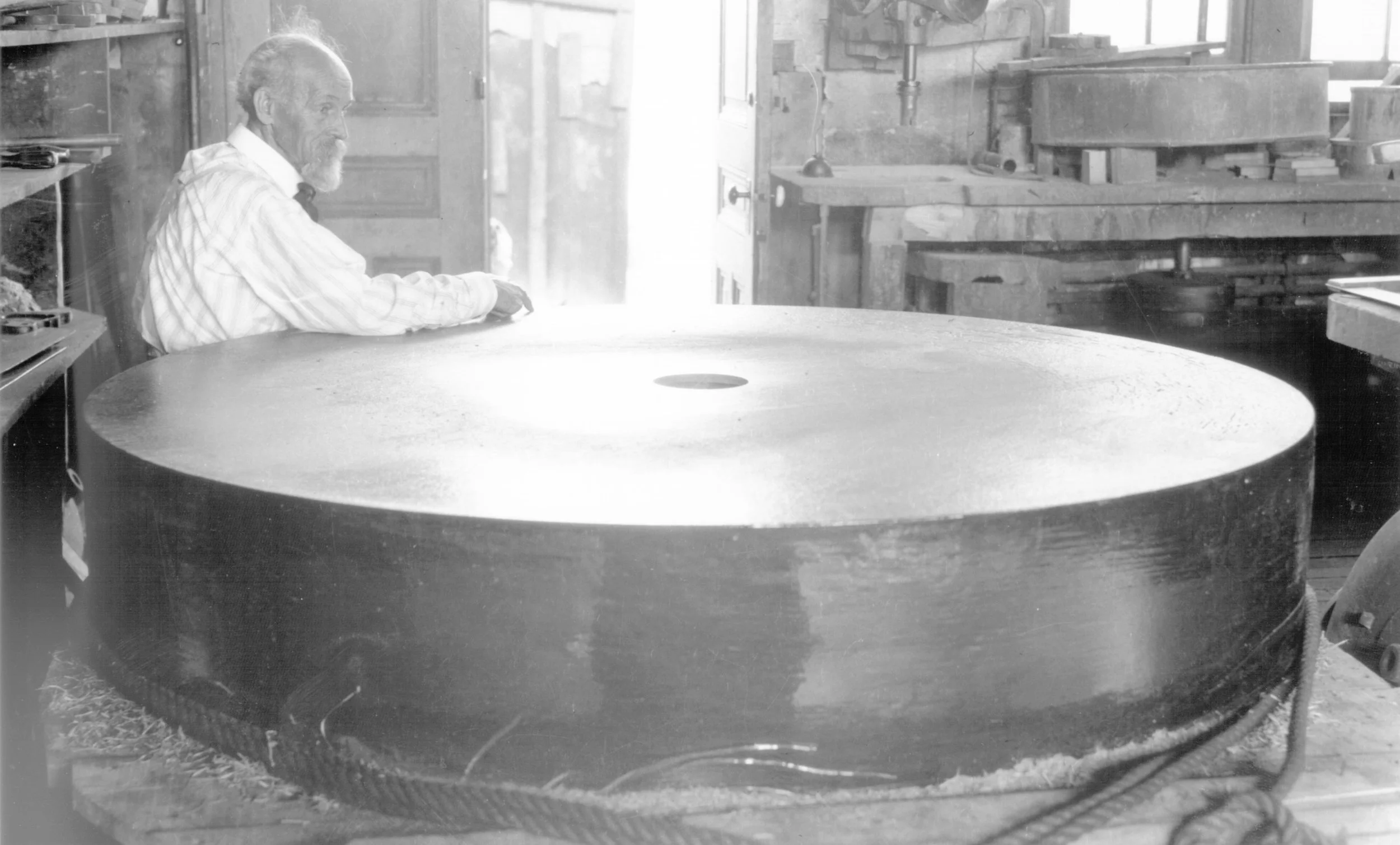 An elderly man sitting next to a large circular metal disc in a workshop.