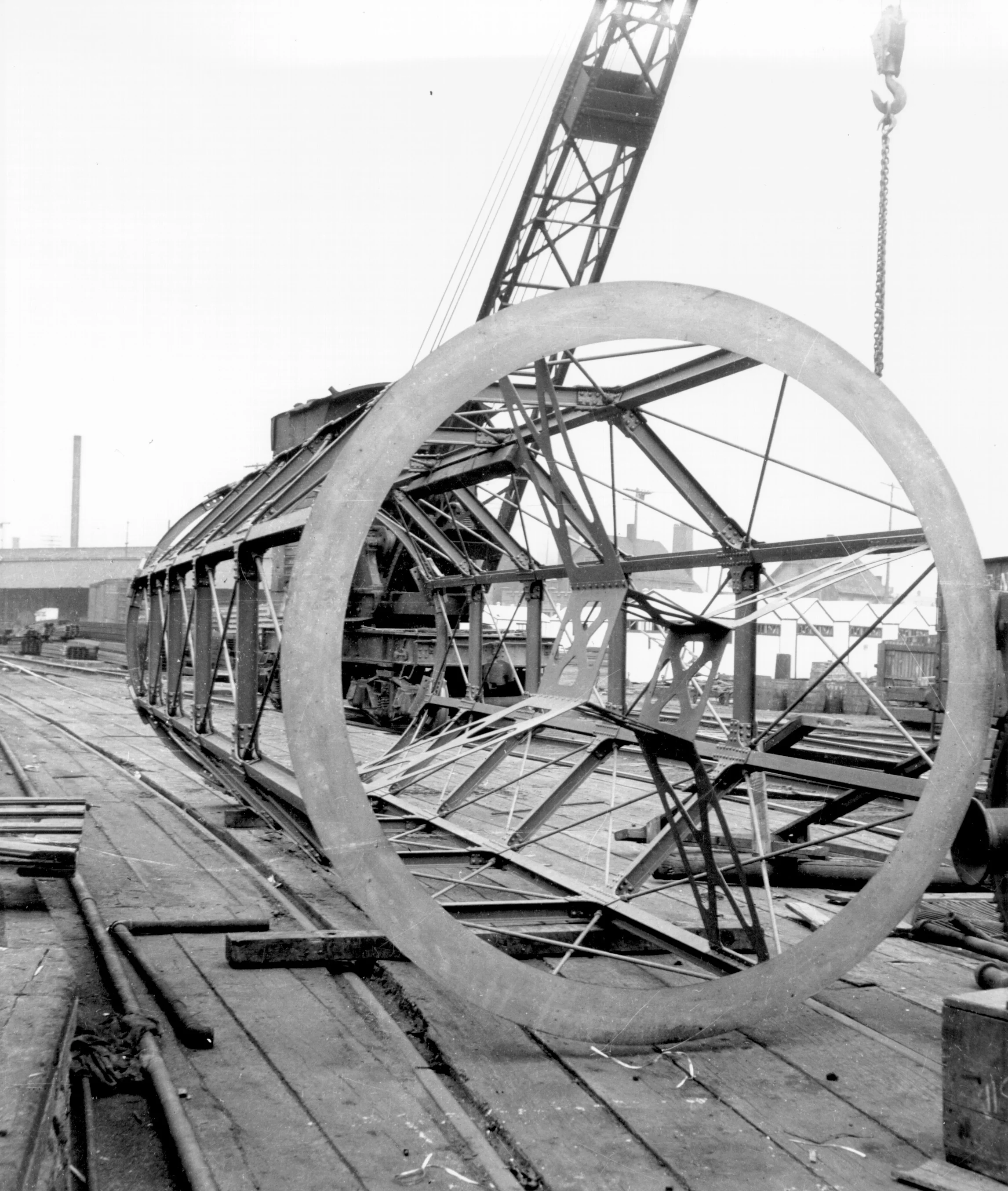 A large metal structure resembling an industrial wheel or framework lying on its side, with a crane and industrial buildings in the background.