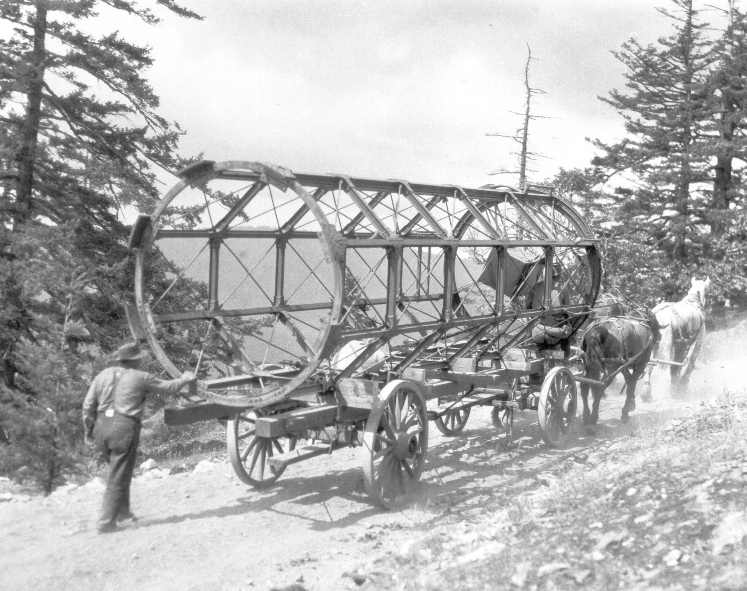 Historical black and white photo of a large, cylindrical framework being transported on a horse-drawn wagon with a person guiding it, surrounded by trees on a dirt path.