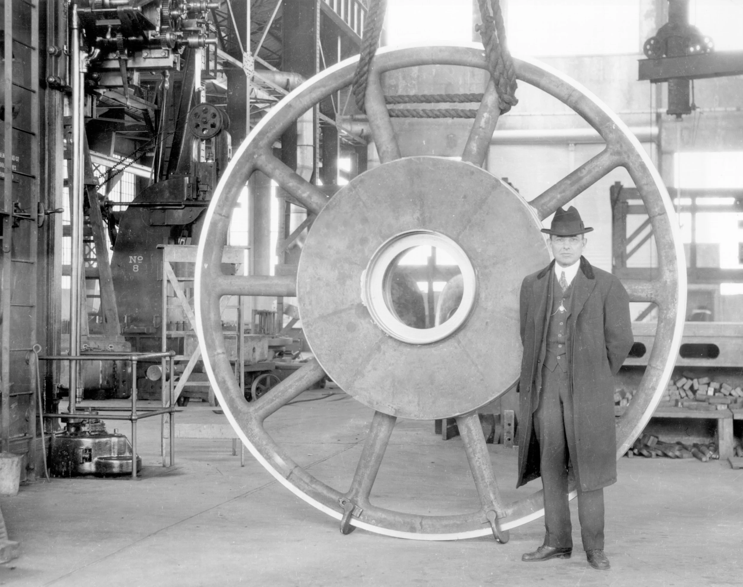 A man wearing a suit and hat stands next to a large, industrial-sized wheel in a factory setting. The wheel is mounted vertically and held in place by machinery. The background shows various industrial equipment and structures, typical of an early 20
