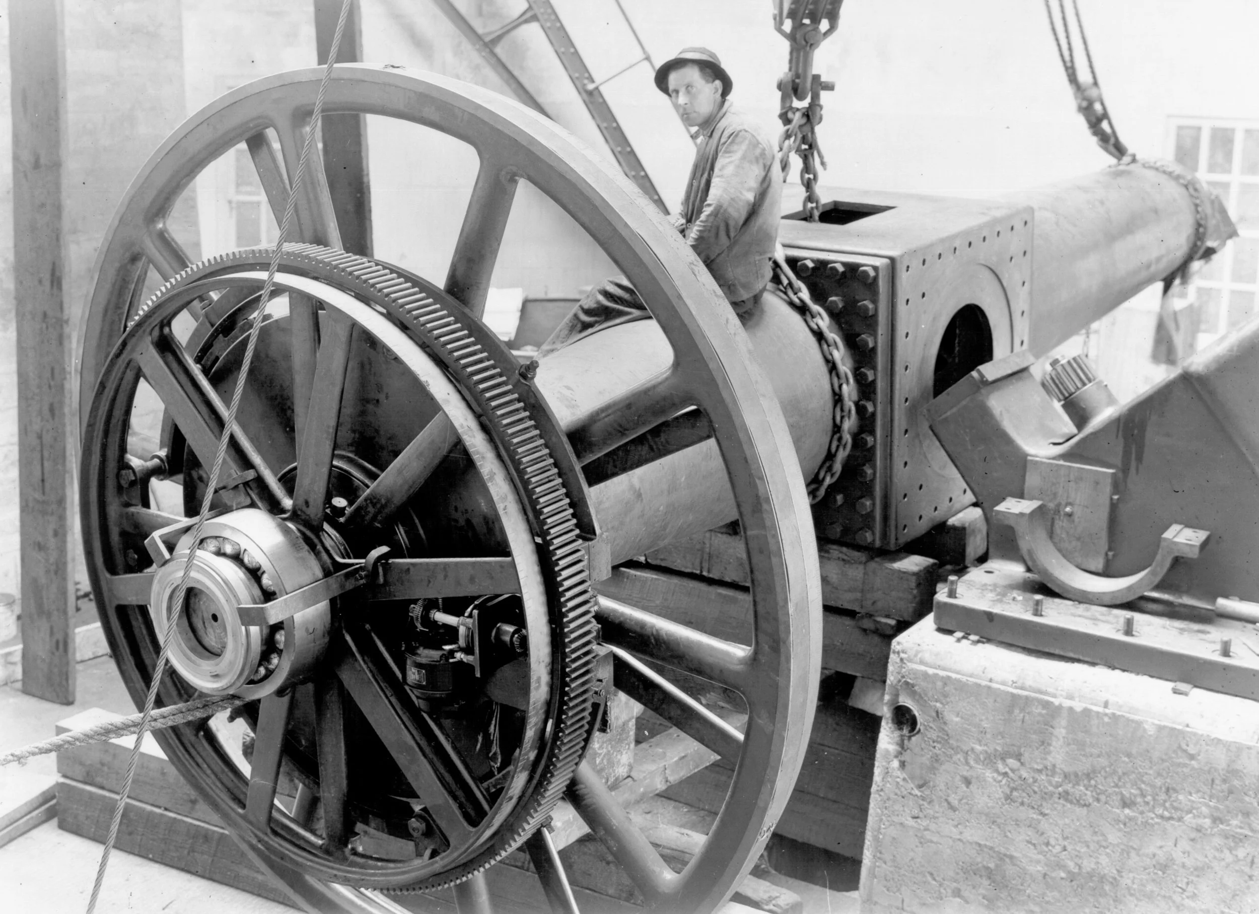Large industrial machinery with gears and a man wearing a hat and work clothes seated on the machinery.