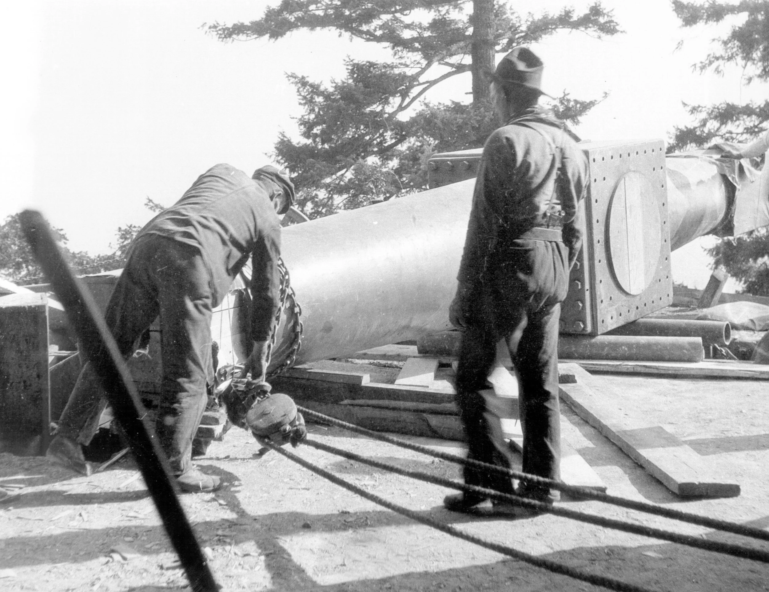 Two workers securing a large metal structure with ropes and equipment outdoors, surrounded by trees.