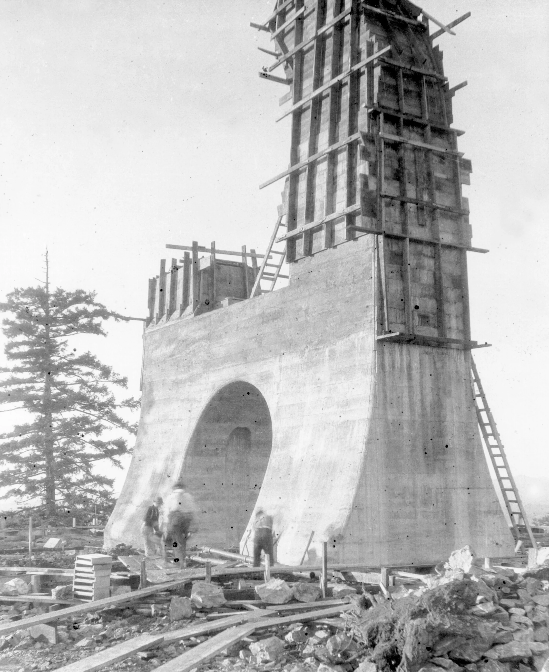 Construction workers building a concrete structure with wooden scaffolding on a rocky terrain. A large arch is part of the structure. There is a tree in the background.