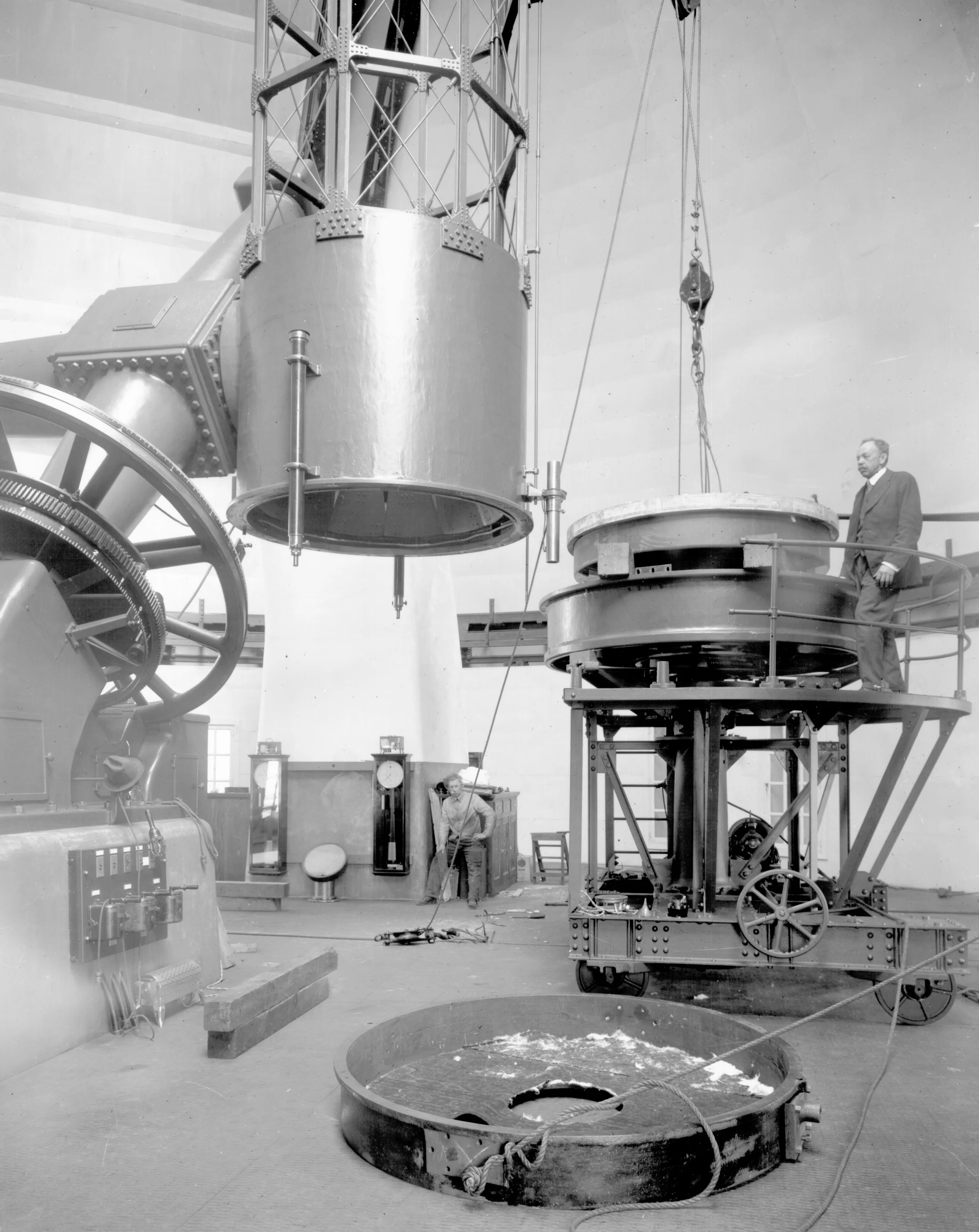 Black and white historical photo of scientists working with large machinery in a laboratory. Two men are visible, one in the foreground and one in the background, manipulating industrial apparatus.