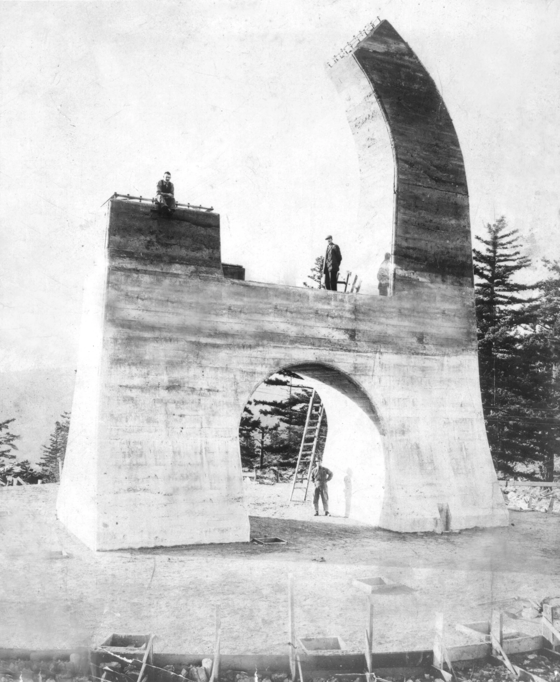 Historic construction site showing an unfinished concrete arch structure with workers and a ladder. Trees are visible in the background.