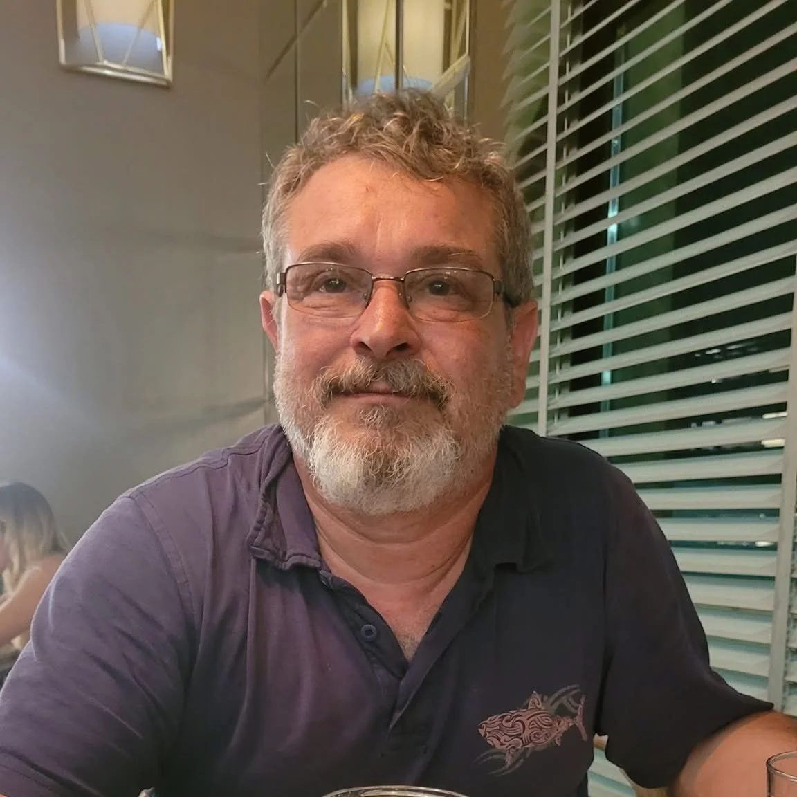 Man with glasses and beard sitting indoors next to window blinds.