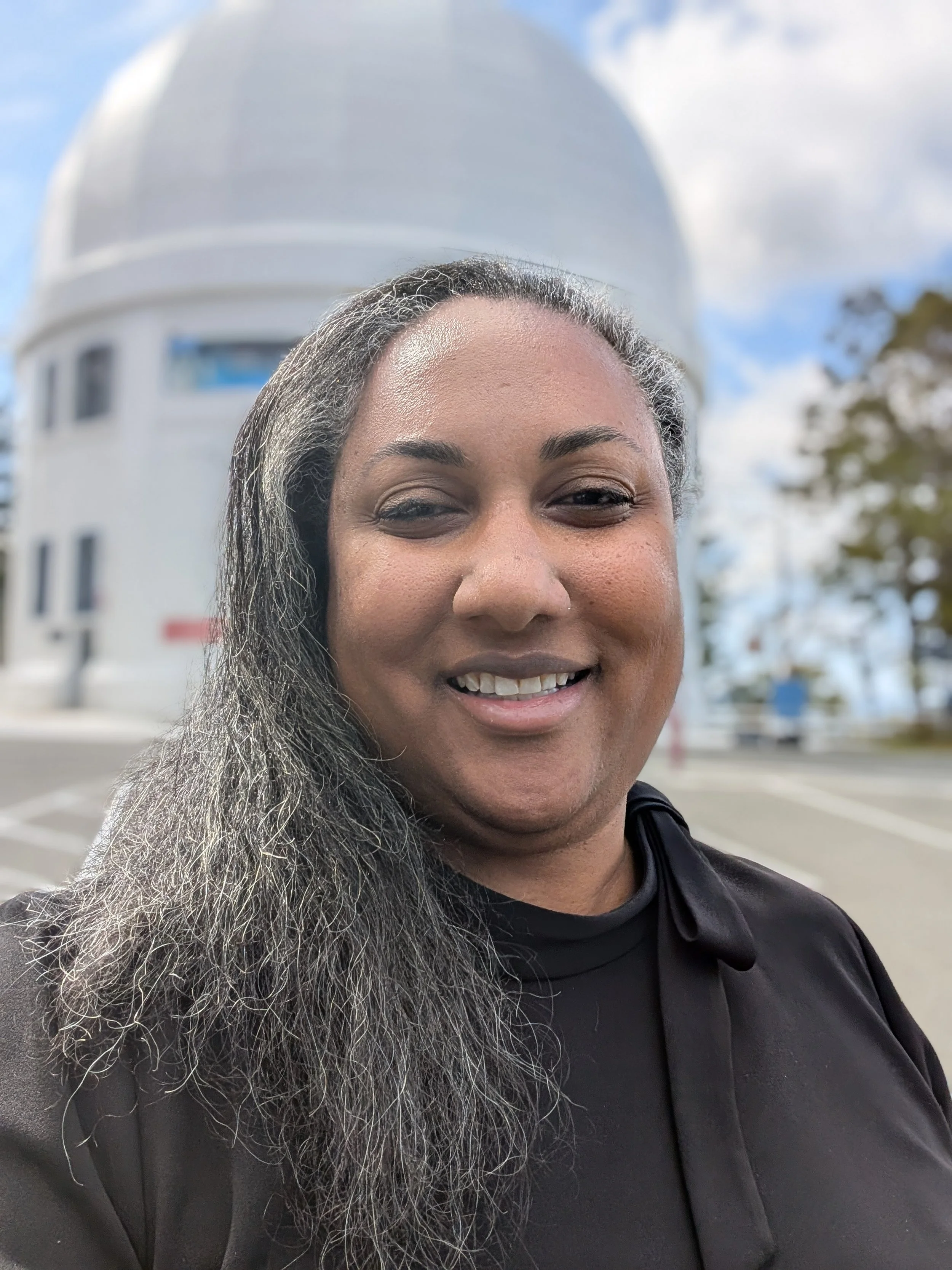 Close-up of a smiling woman with gray hair and a black outfit, standing in front of a white domed observatory with a blue sky and clouds in the background.