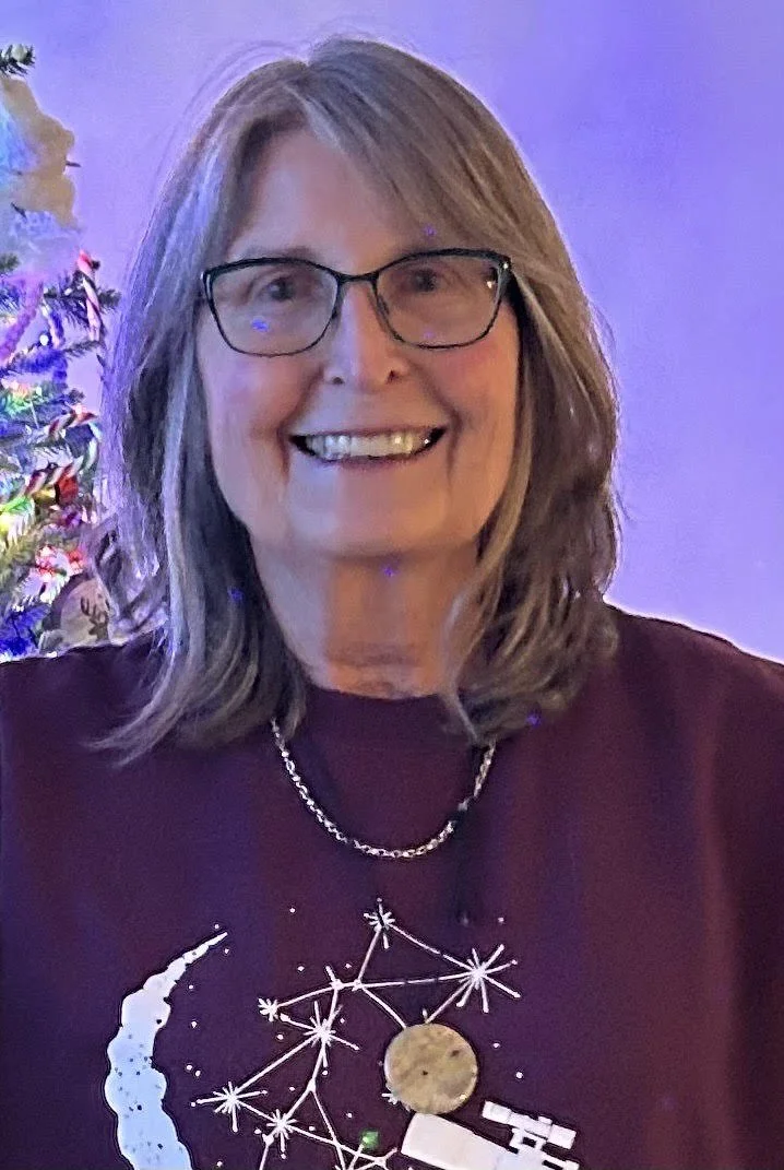 Smiling person wearing glasses and a festive sweater, standing near a decorated Christmas tree with blue lighting.