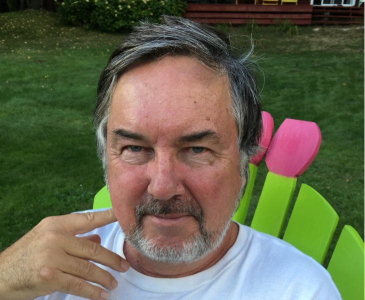A man with gray hair and a beard sitting on a colorful chair outdoors on green grass.