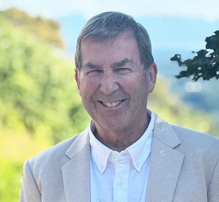A smiling middle-aged man wearing a light beige blazer and white shirt outdoors with a blurred natural background.