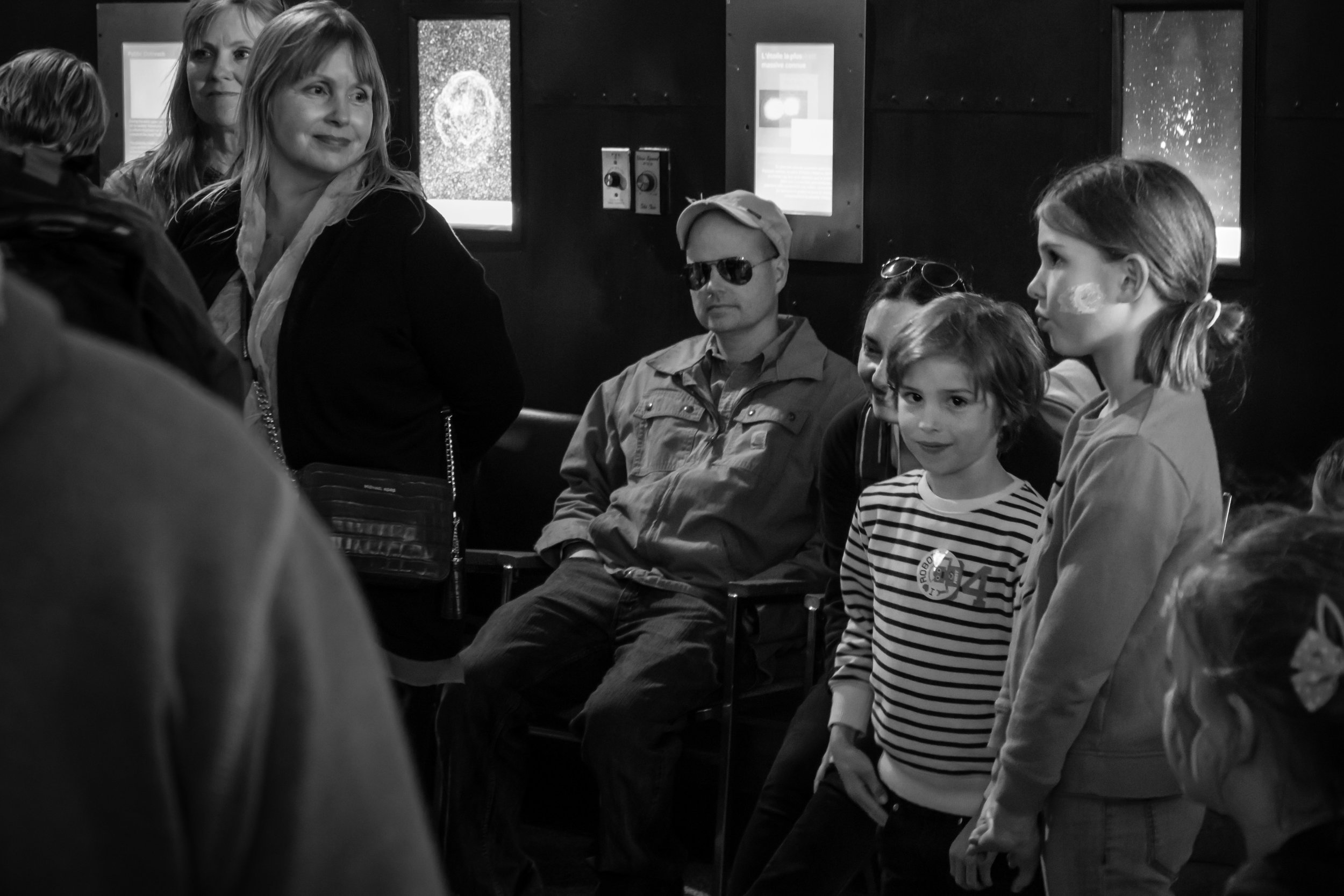 Black and white photo of a group of children and two women, one with blonde hair and another with glasses, inside a dimly lit room. The children are sitting and standing, wearing casual clothes, and some have stickers on their faces. The background shows some windows and informational posters.