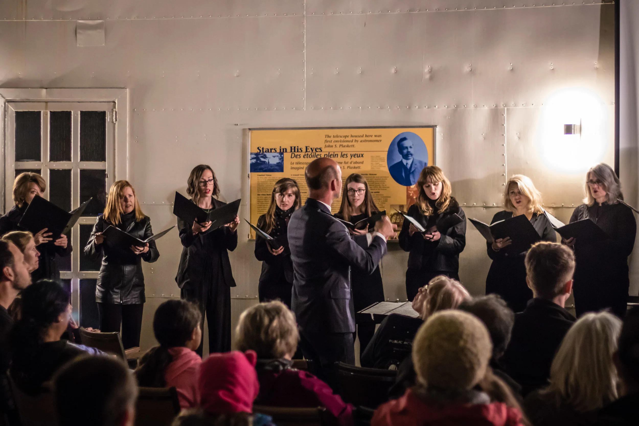 A choir of women singing with sheet music, conducted by a man in a suit, in an indoor setting with an audience watching. There is a yellow informational poster about the history of a telescope behind them.