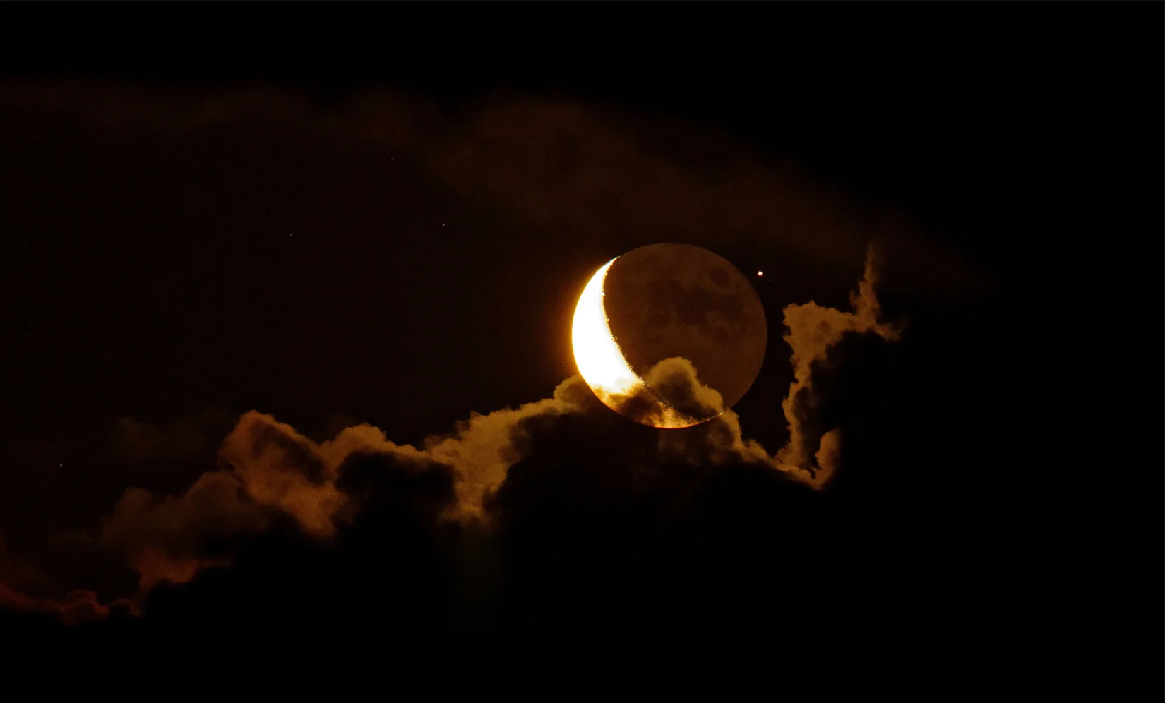 A lunar eclipse captures the moon with a partial shadow causing a crescent-shaped bright outline against the dark night sky, with some clouds illuminated around the moon.