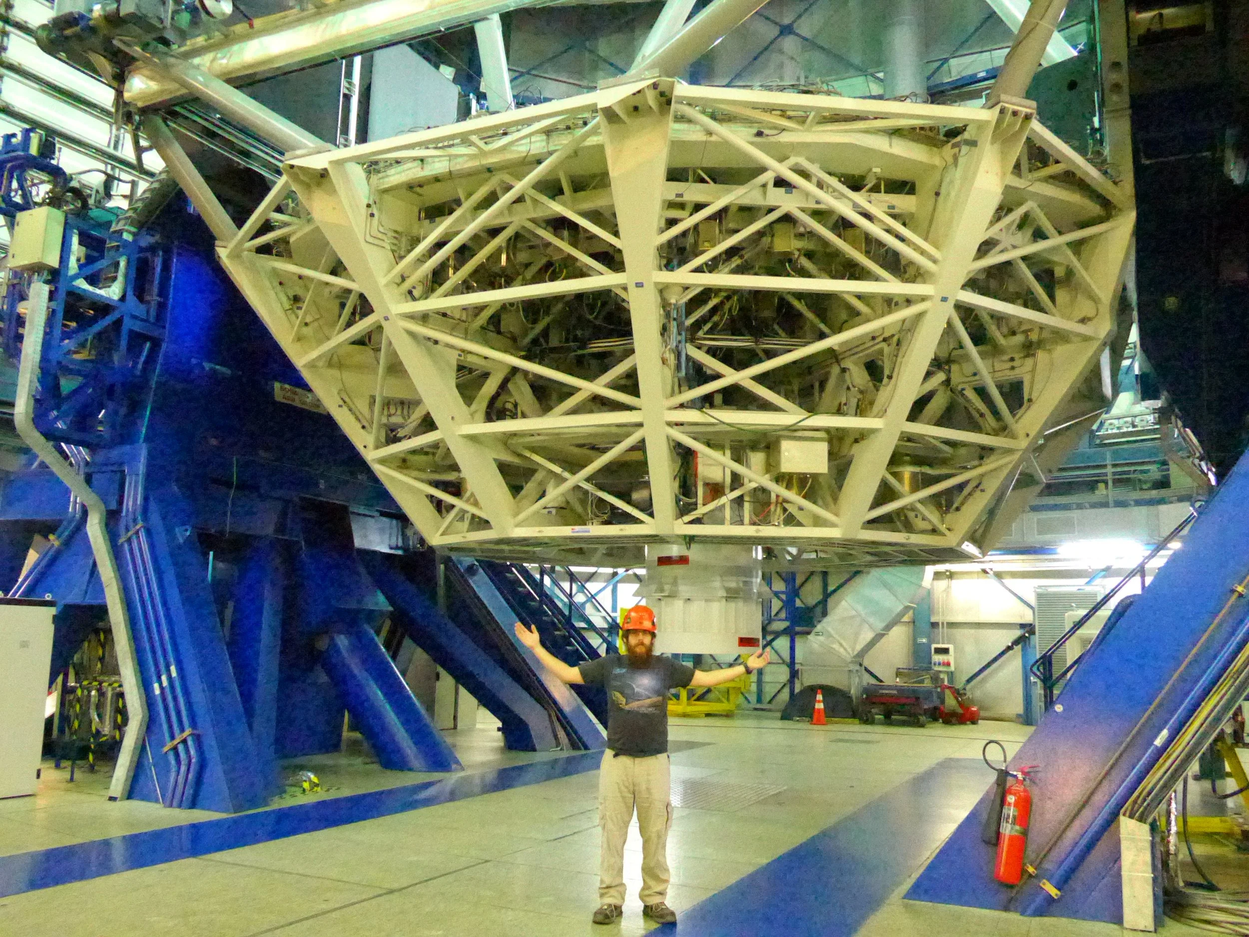 A man wearing a red hard hat and grey pants standing inside a large industrial or scientific facility, with a complex structure of metal framework and large blue and white components behind him.