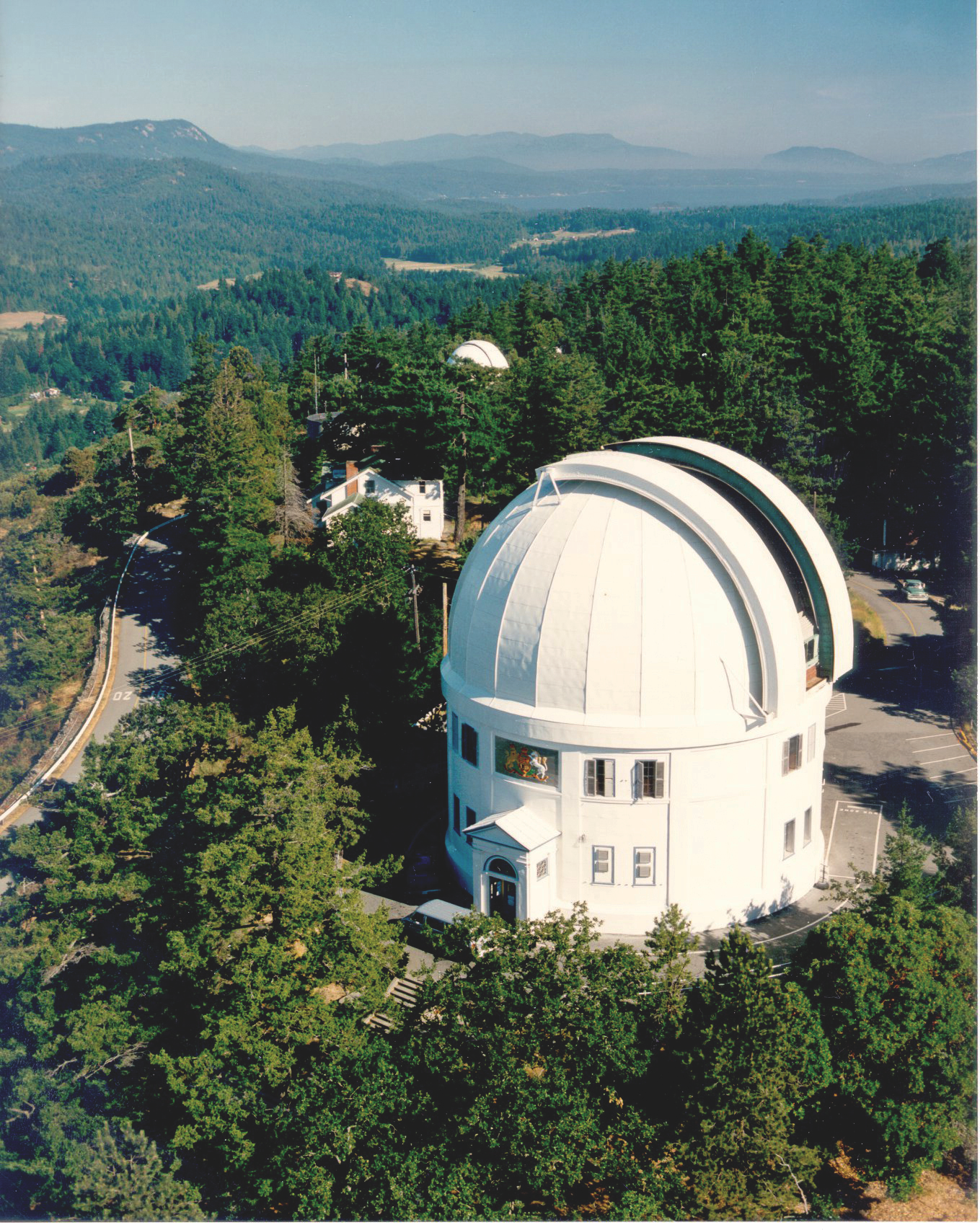 The Dominion Astrophysical Observatory seen from the air, with a white dome-shaped roof situated among tall green trees on a hillside. It offers a view of a scenic landscape with forested hills and mountains in the distance under a clear sky.