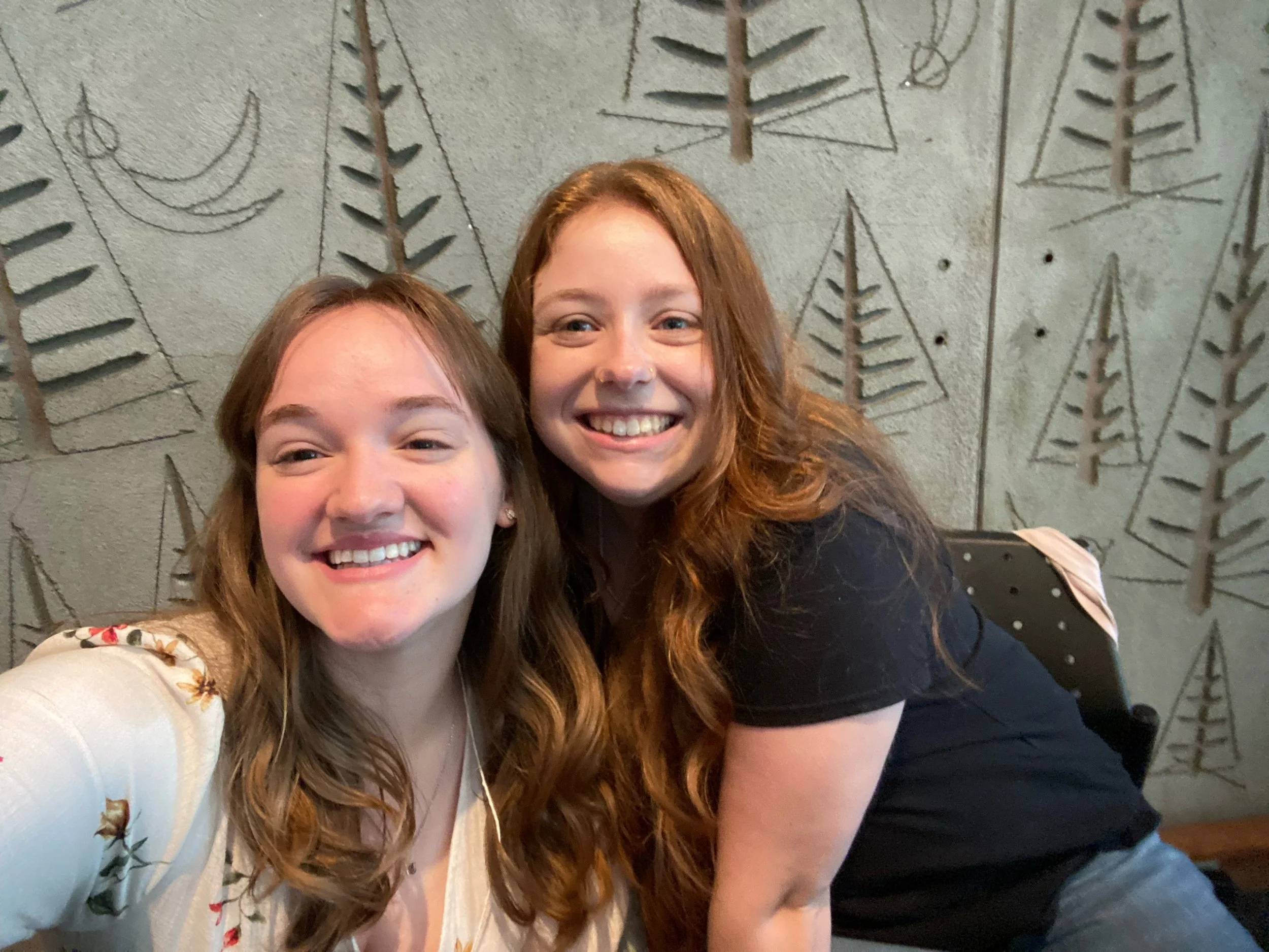 Two young women smiling and taking a selfie together in front of a decorative wall with engraved geometric and botanical patterns.