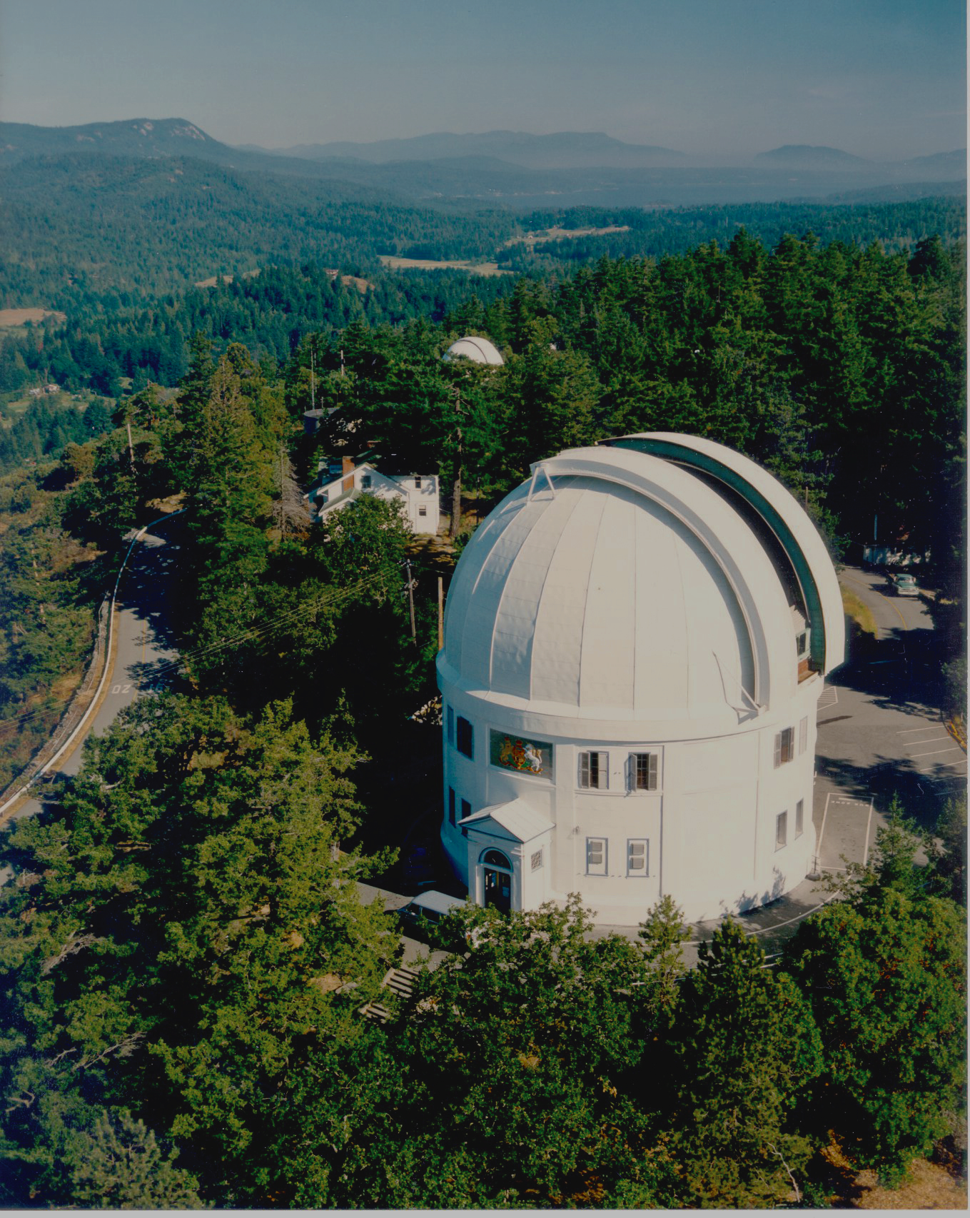 A white observatory with a domed roof in a forested area, with mountains and a clear sky in the background.