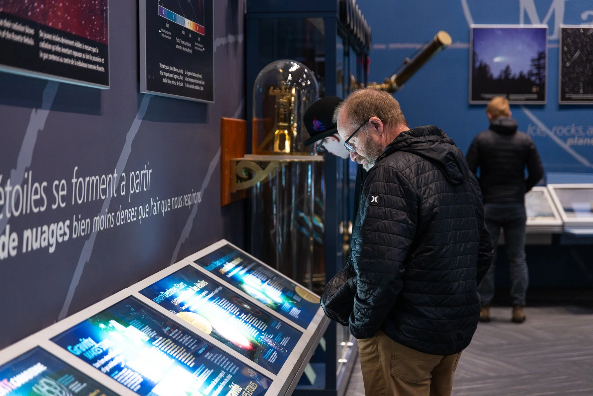 A man with glasses, a beard, and a black jacket is looking at an informational display with multiple digital screens at a science or space exhibit. In the background, another person is walking by, and there are posters and a large telescope.