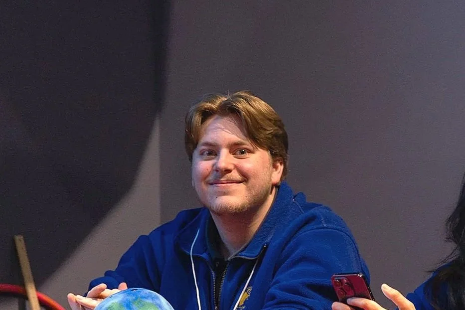 A young man with brown hair and a beard smiling at the camera, sitting at a table with a globe and a smartphone in his hands, wearing a blue jacket.