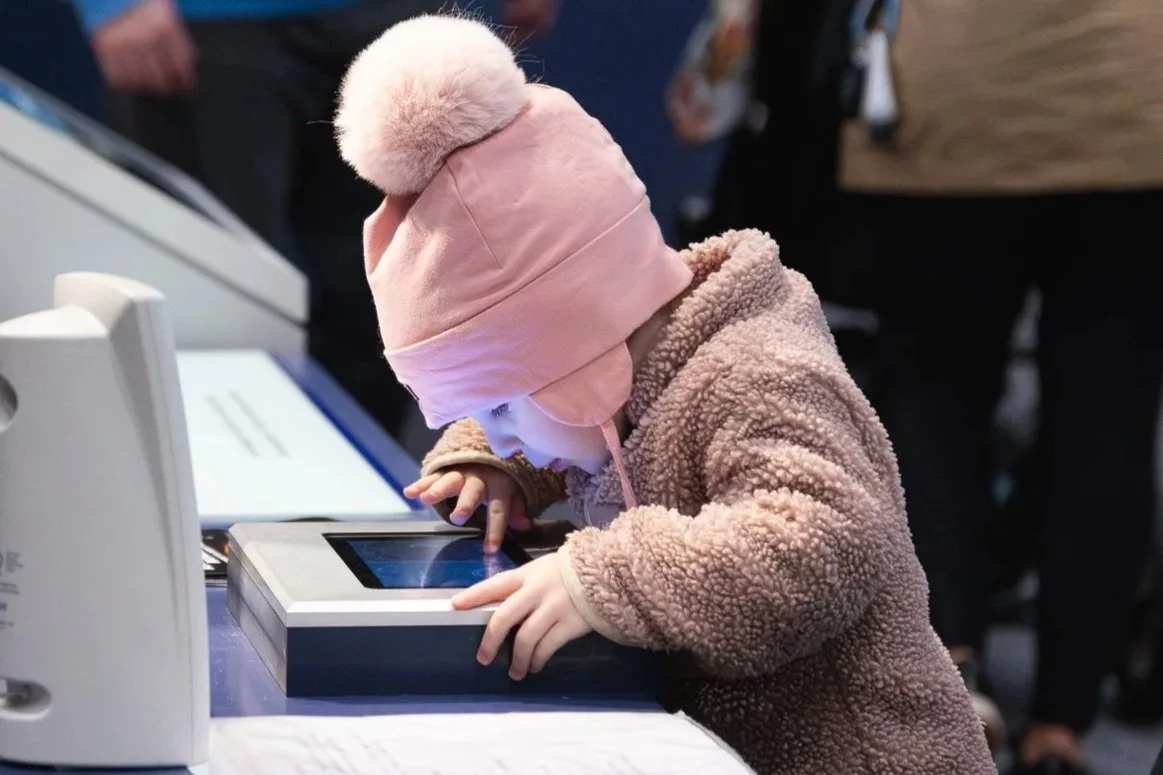 Young girl in pink fleece coat and matching pink beanie with pom-pom using a touchscreen device at a table.