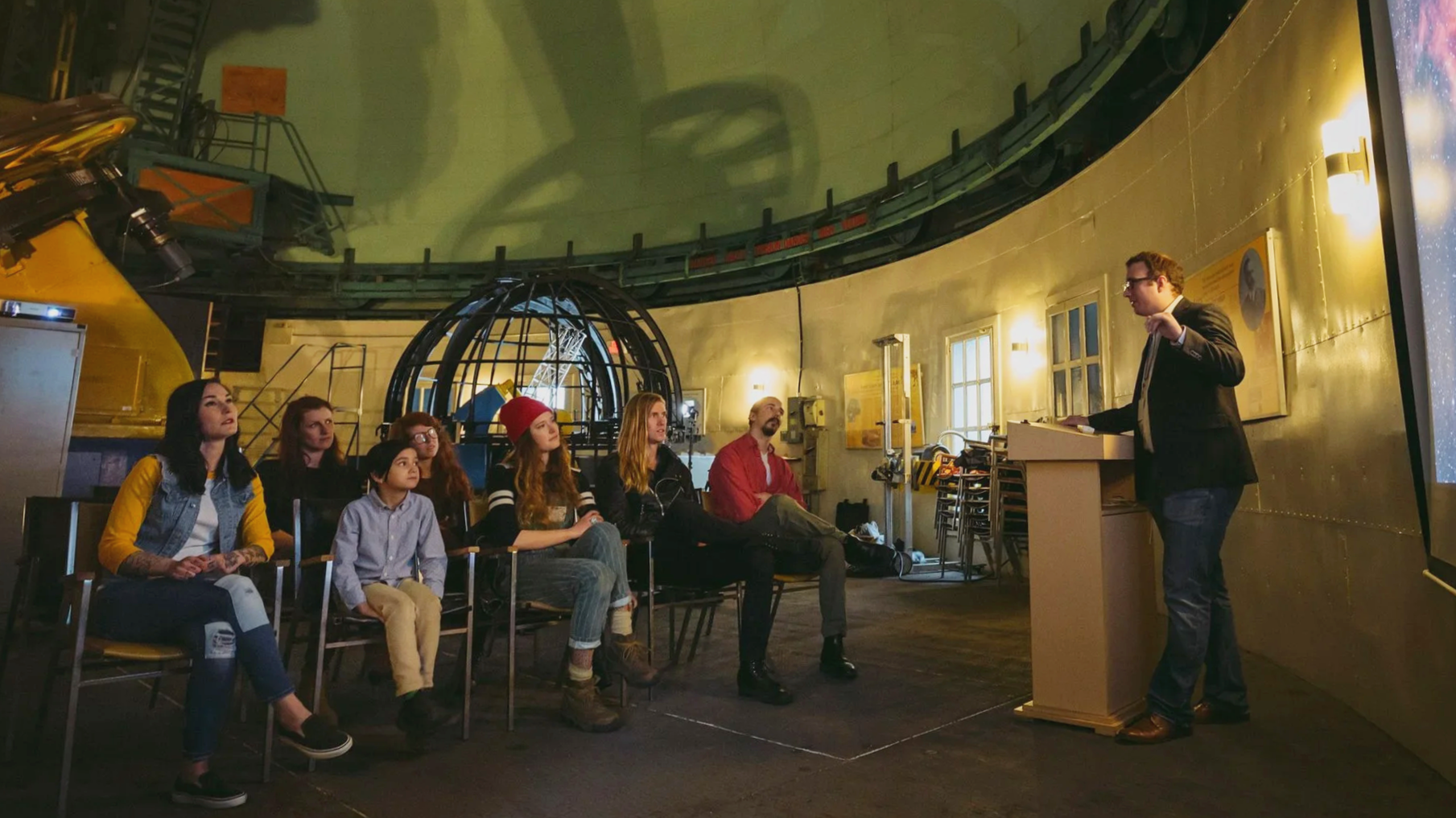 A man in a suit giving a lecture to a group of diverse young people seated in chairs inside a planetarium with a domed ceiling and astronomical equipment.