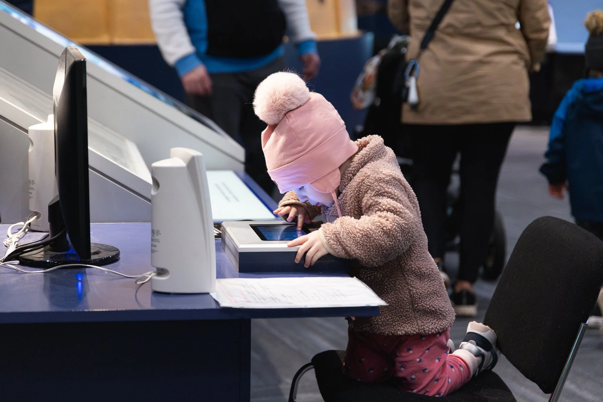 A young girl wearing a pink fleece jacket, a pink hat with a pompom, and pink pants with colorful hearts, is kneeling on the floor and touching a touchscreen monitor set up on a table. Other people are standing in the background, blurred.