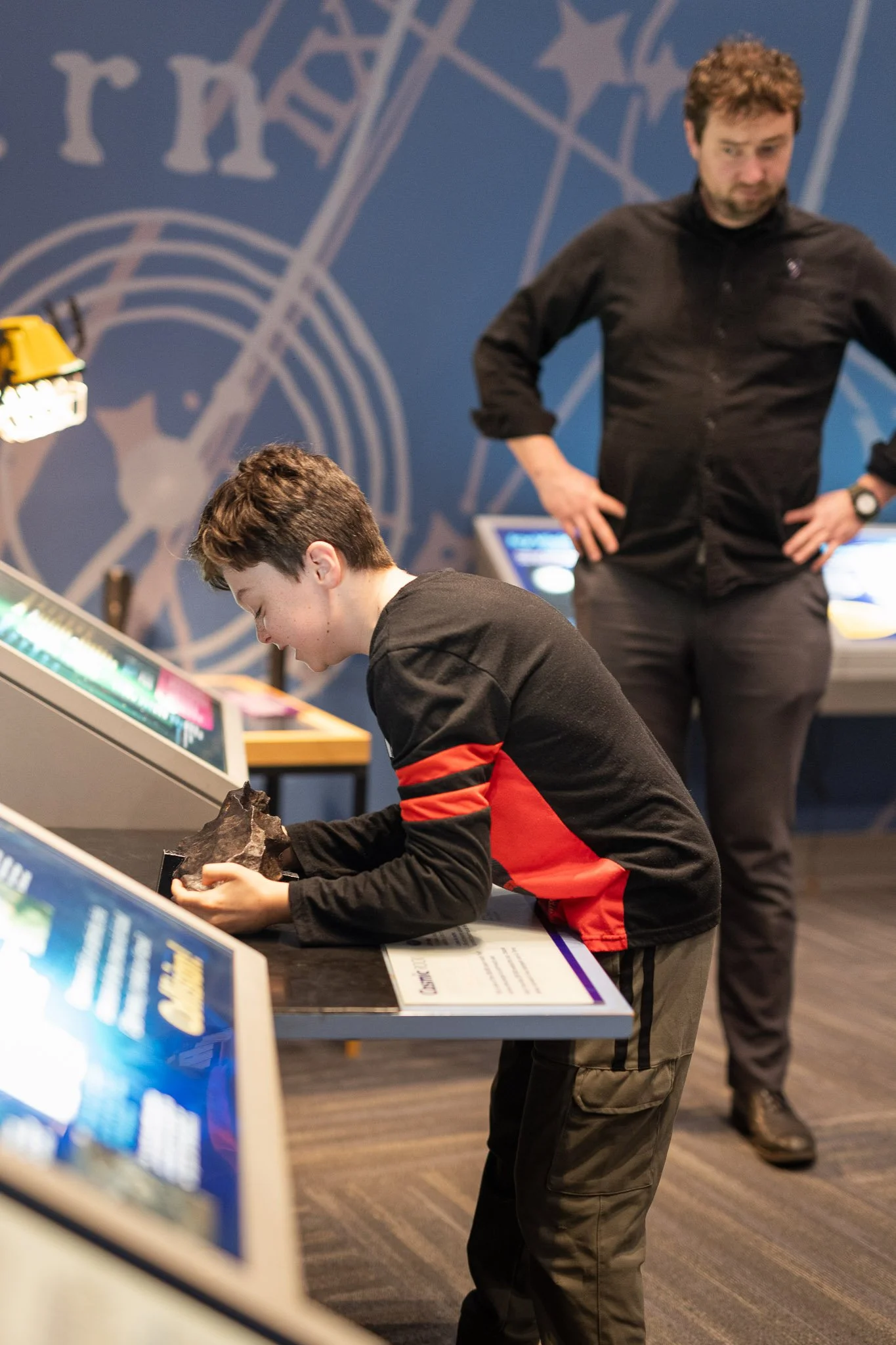 A boy in a black and red jacket is examining a rock at an educational exhibit, with a man in black standing behind him. The background shows a blue wall with a large white gear and clock design, indicating a science or technology setting.