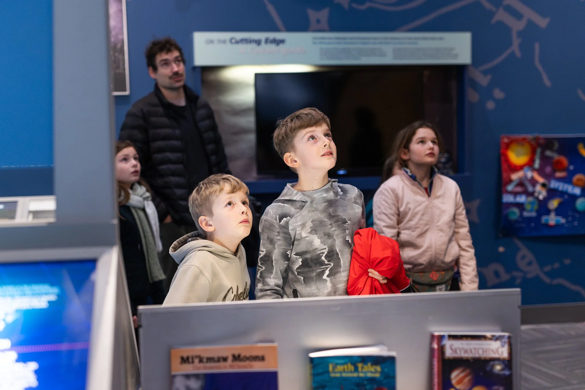 A group of children and one adult are inside a science or discovery center, looking upwards with curiosity. They are standing near an exhibit or display, with a blue background and some books visible in the foreground.