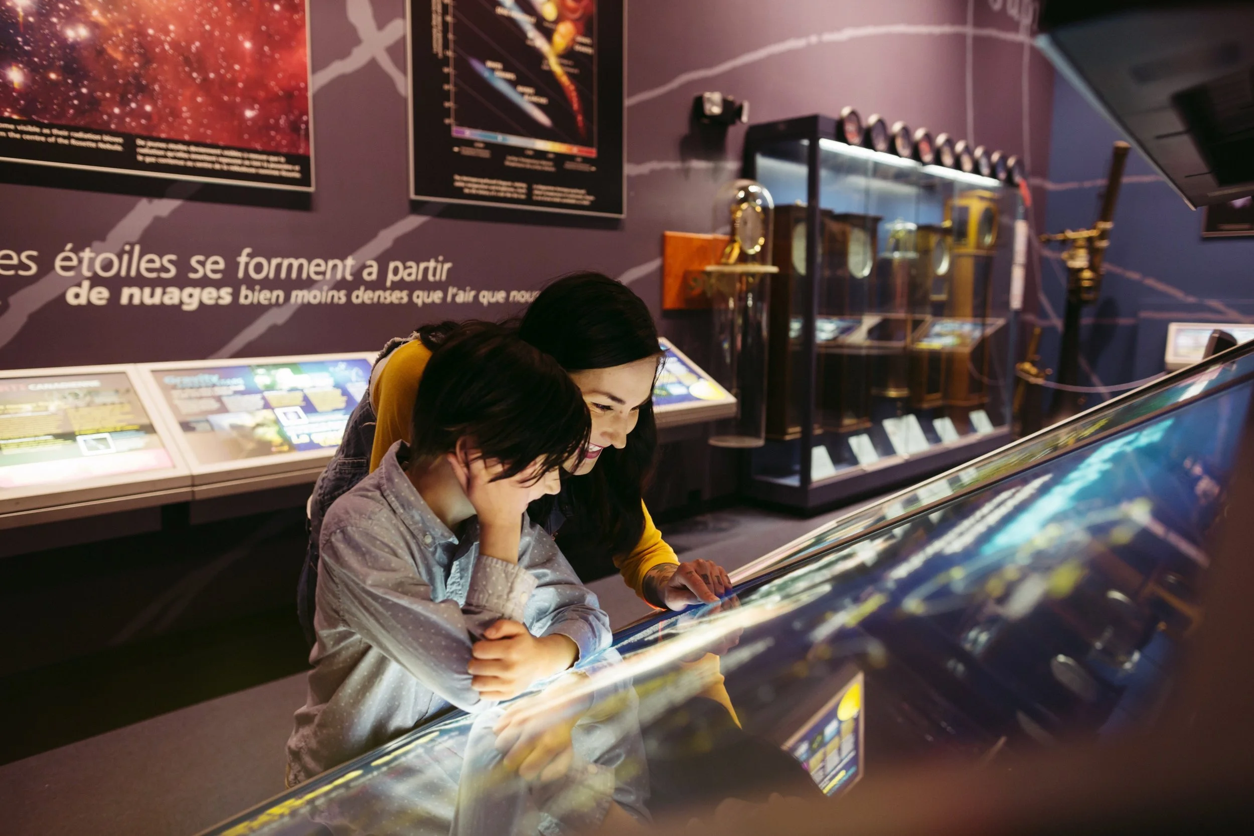 Two children and an adult looking at a star map exhibit in a science museum, with educational posters and display cases in the background.