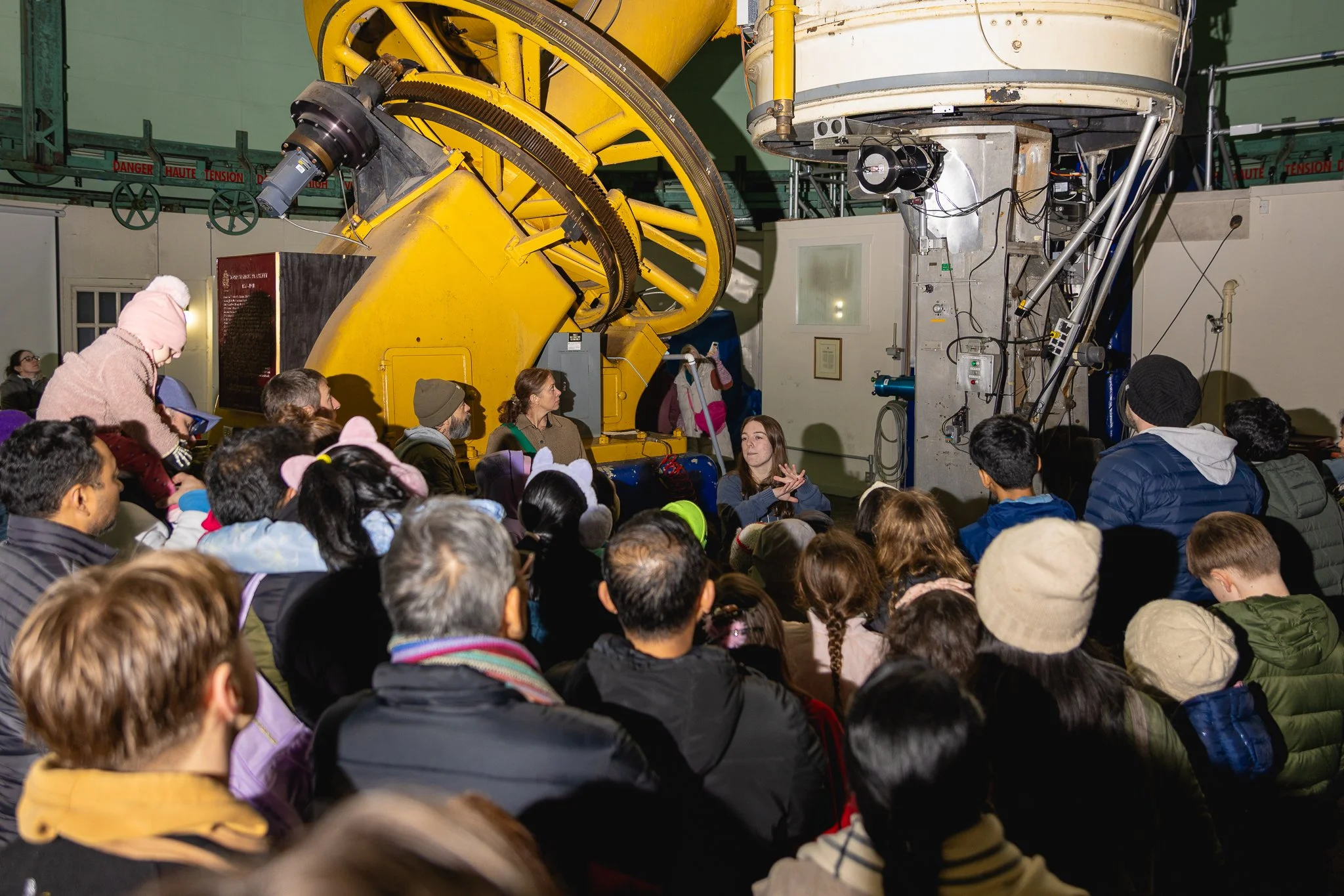 Group of people gathered around a large yellow space telescope, observing and listening to a guide explaining the telescope's features.
