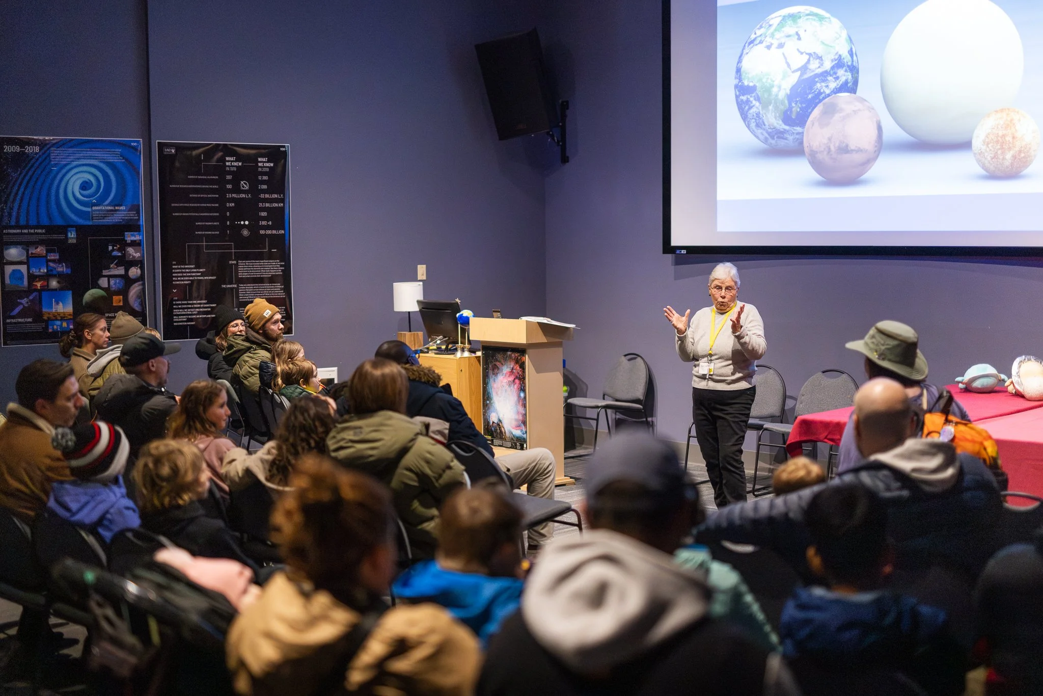 An elderly woman is giving a presentation in a room filled with seated audience members. Behind her is a large screen displaying images of planets and a poster related to astronomy.