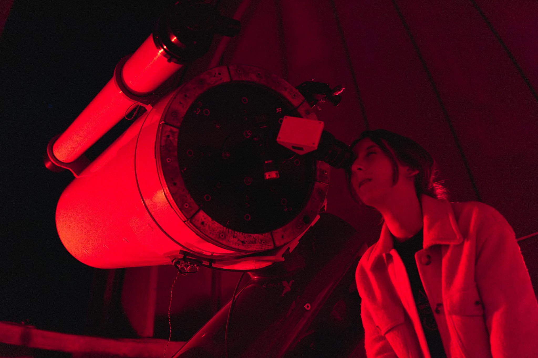 A person working on a large astronomical telescope illuminated by red light, lying on the ground, with various machinery and cables nearby.
