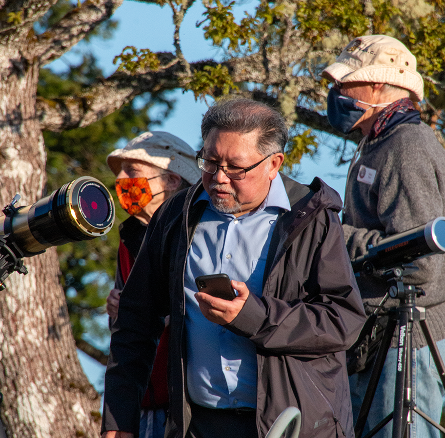 Three people outdoors near a large tree, with one man looking at his phone while two women wear masks and look at a telescope.