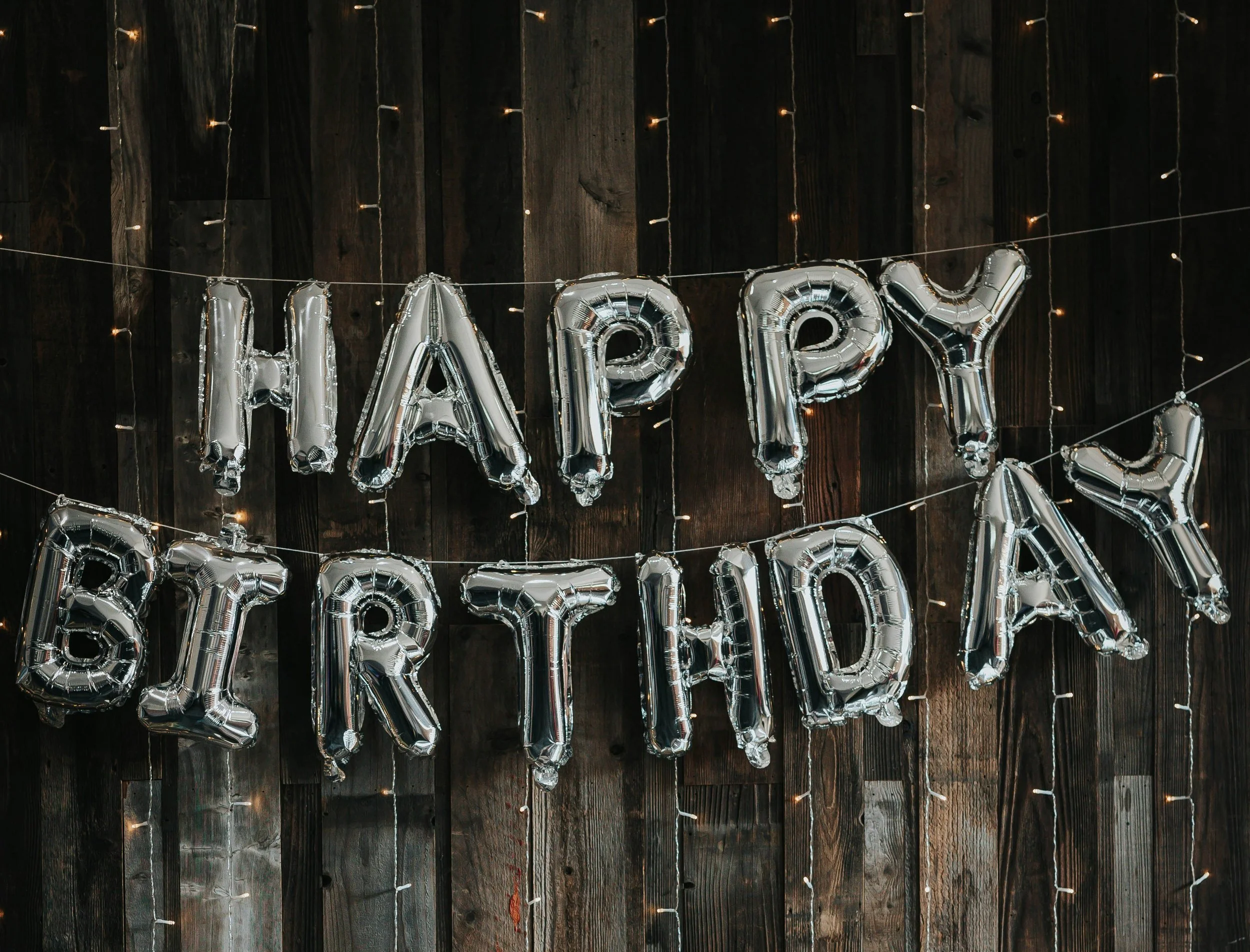 Silver 'Happy Birthday' balloons hanging on a dark wooden wall with string lights.