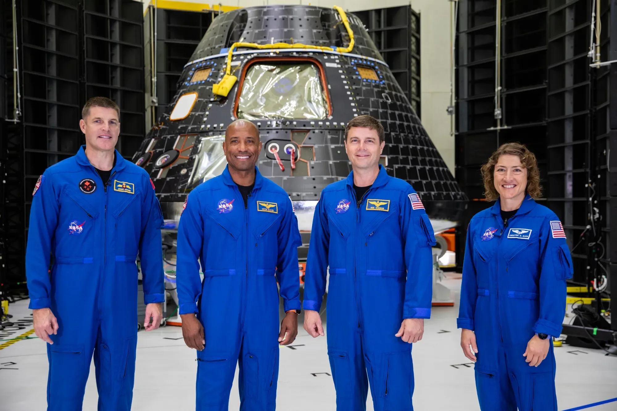 Four NASA scientists in blue jumpsuits standing in front of a spacecraft in a manufacturing or testing facility.