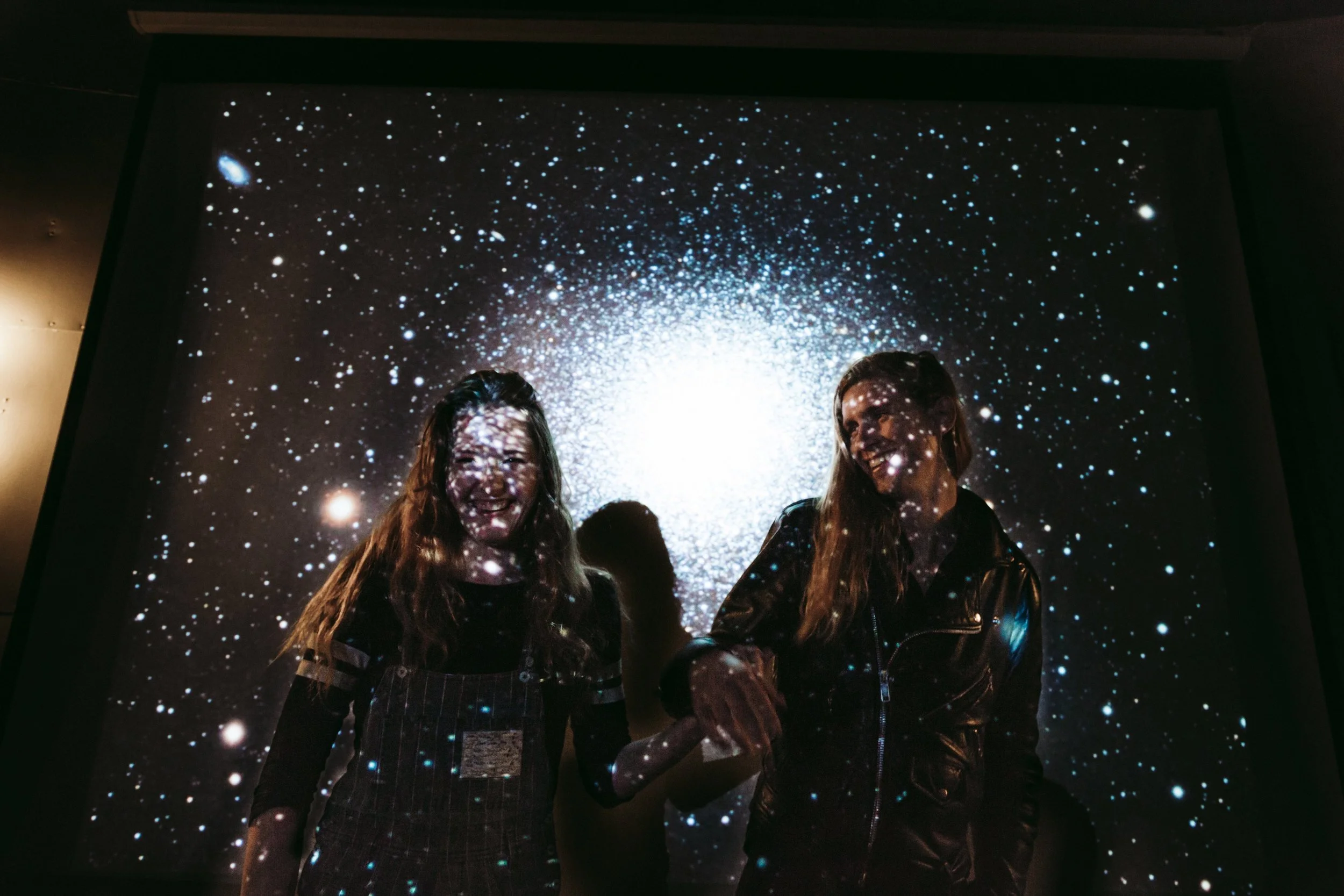 Two women are smiling and holding hands as they stand in front of a large screen projecting an image of stars and galaxies, creating a cosmic background.