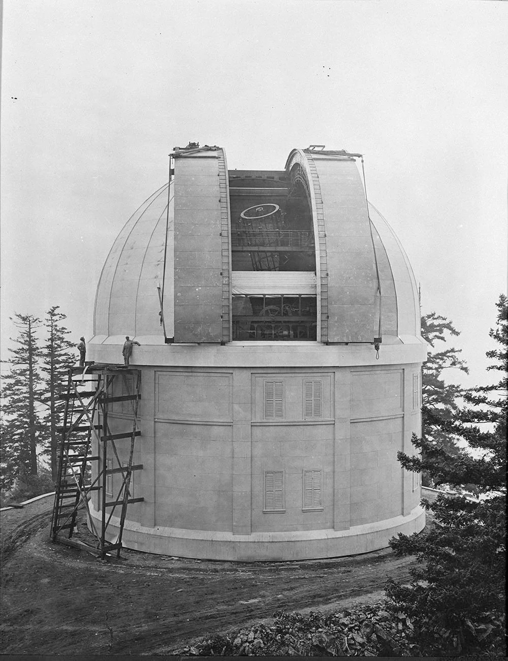 The Griffith Observatory under construction, featuring a large domed structure with a partially assembled telescope inside and workers on scaffolding, surrounded by trees.
