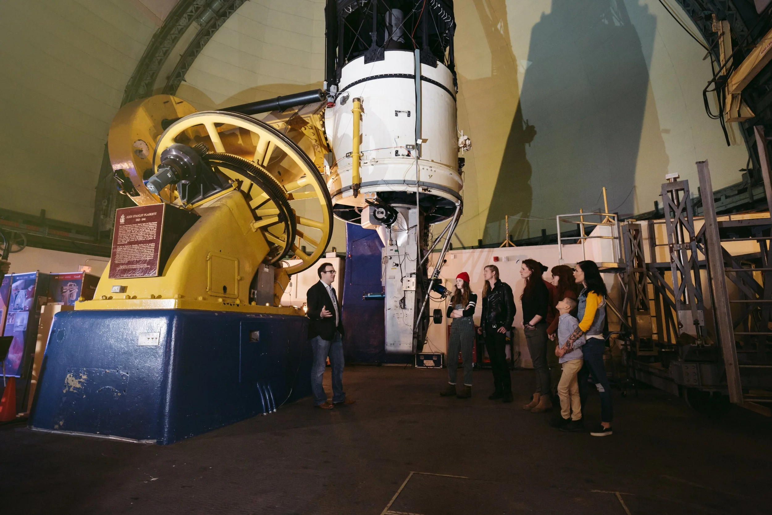 A group of people observing a large spacecraft inside a planetarium or museum setting, with a museum staff member explaining the exhibit.
