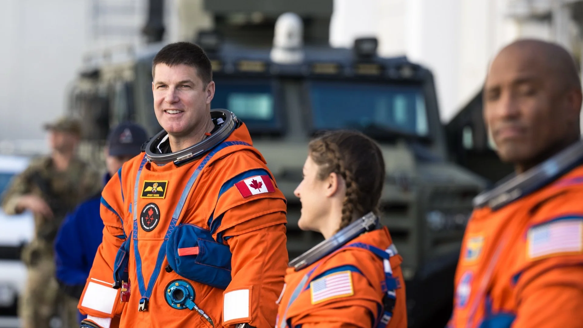 Group of astronauts in orange space suits with Canadian and American flags, standing outdoors near vehicles, smiling and talking.