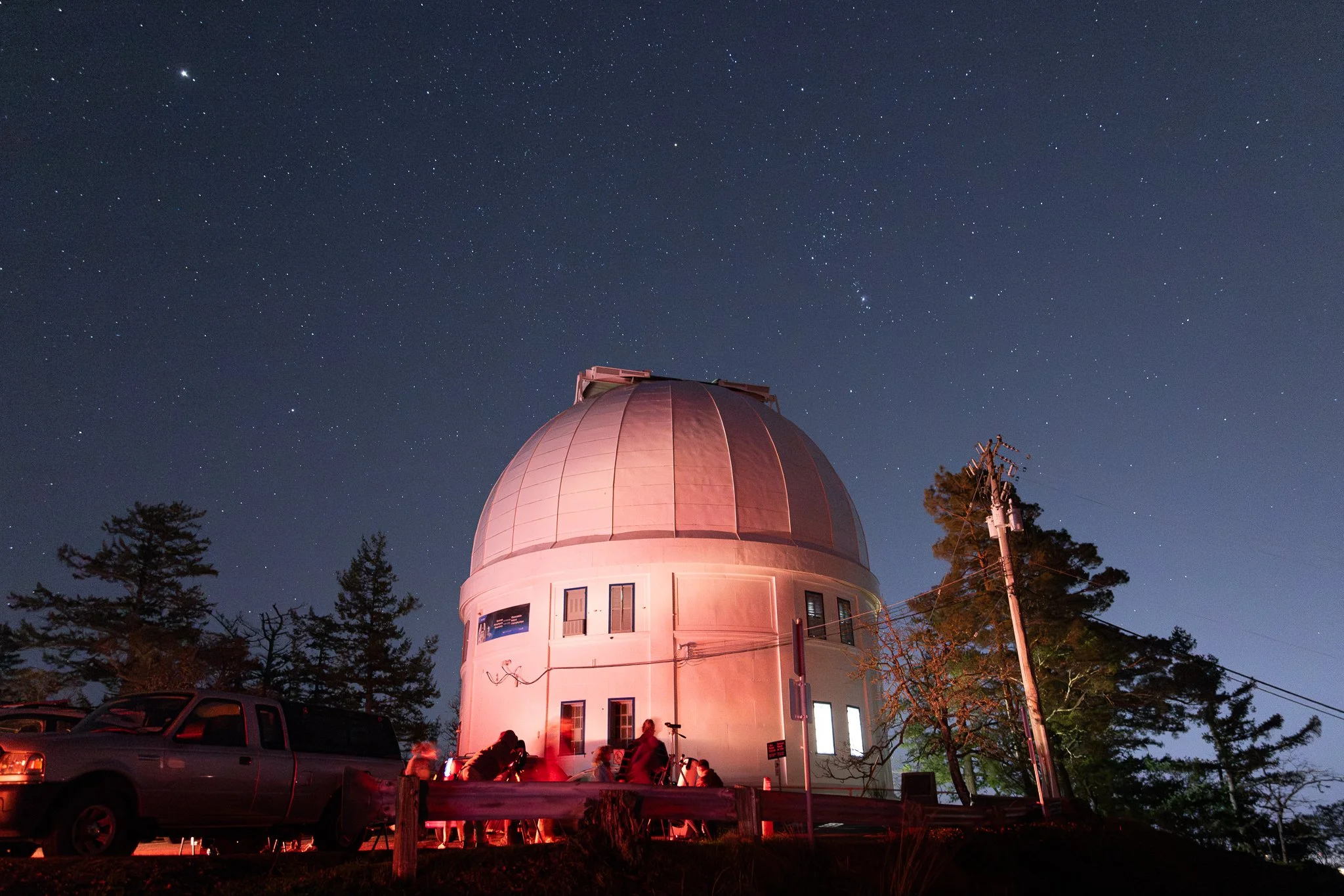 Night sky filled with stars over a white observatory dome, surrounded by trees and parked cars.