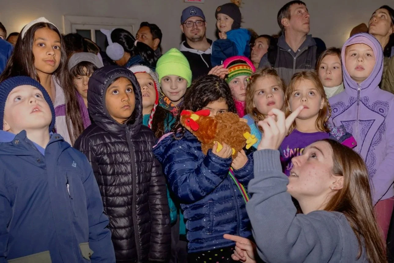 Group of children and adults indoors, some children wearing colorful hats, one girl holding a plush chicken, all looking attentively in the same direction.