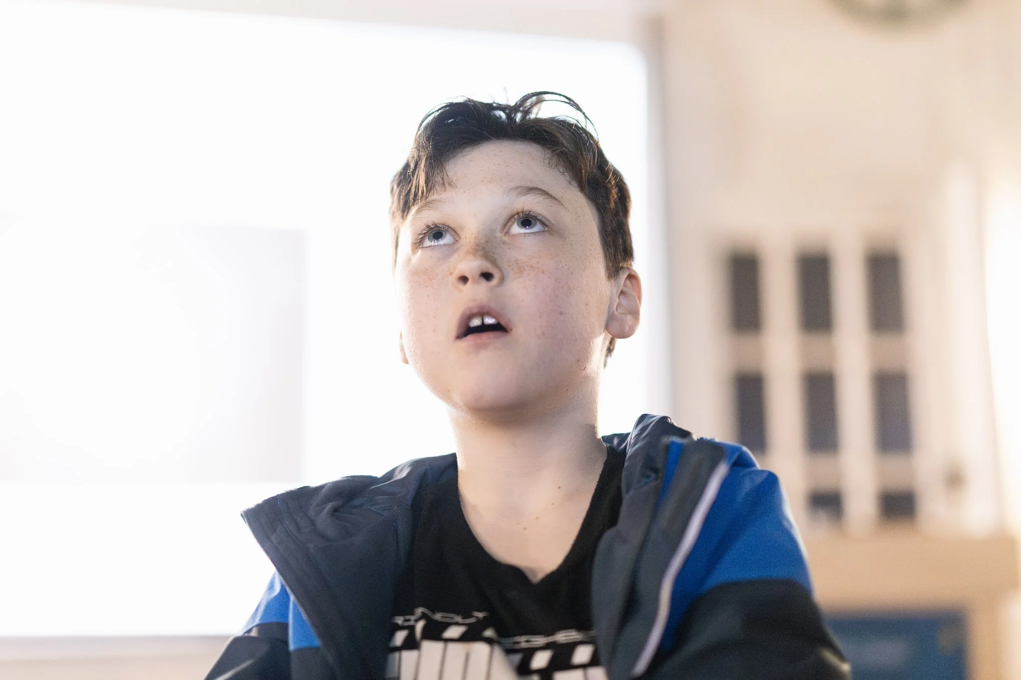 A young boy with light skin, freckles, and short dark hair looking upwards with a surprised or curious expression. He is wearing a black and blue jacket.