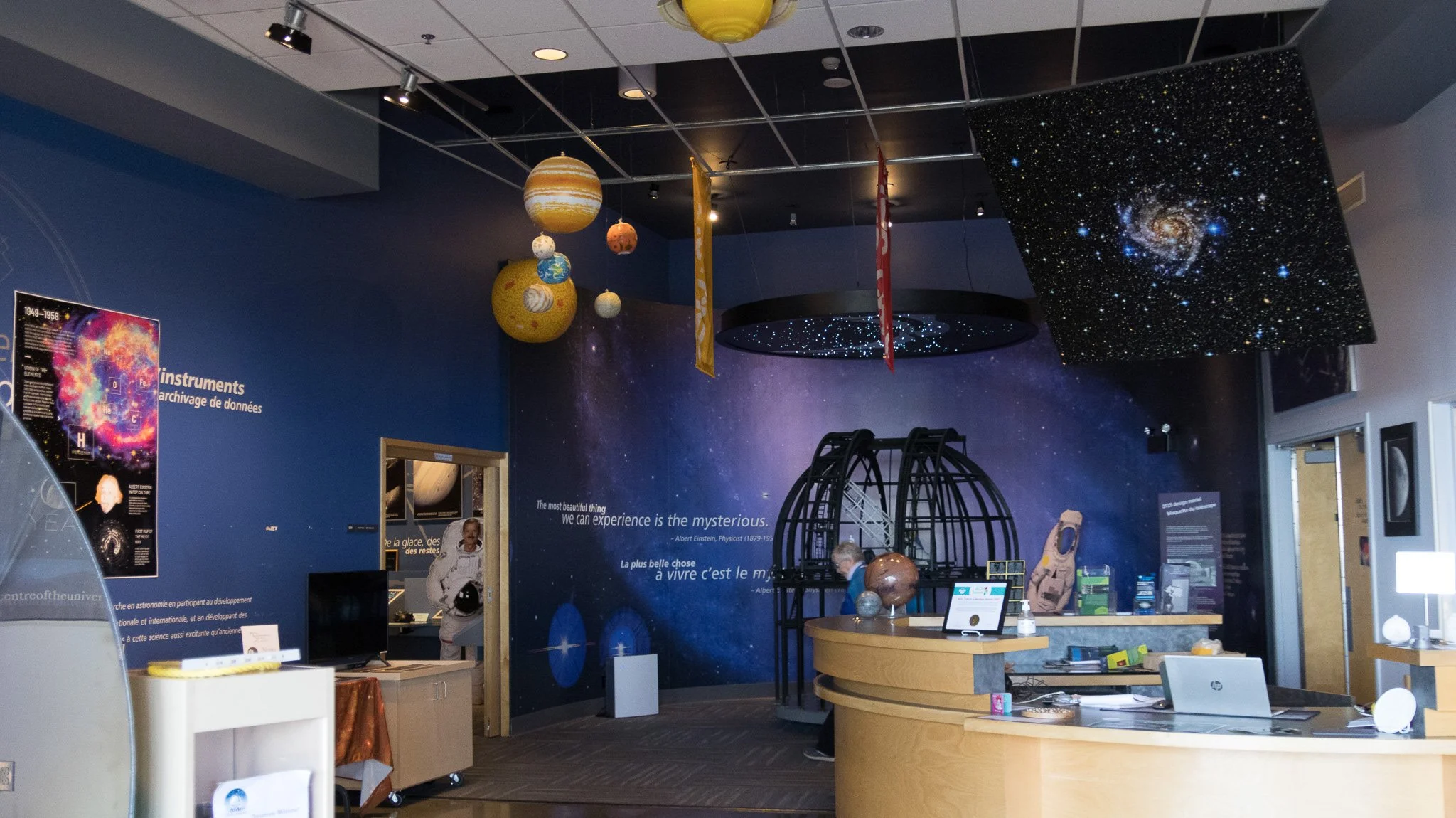 A science museum exhibit featuring a space theme with planetary models hanging from the ceiling, a large wall display of a galaxy, and informational posters about astronomy and the universe.