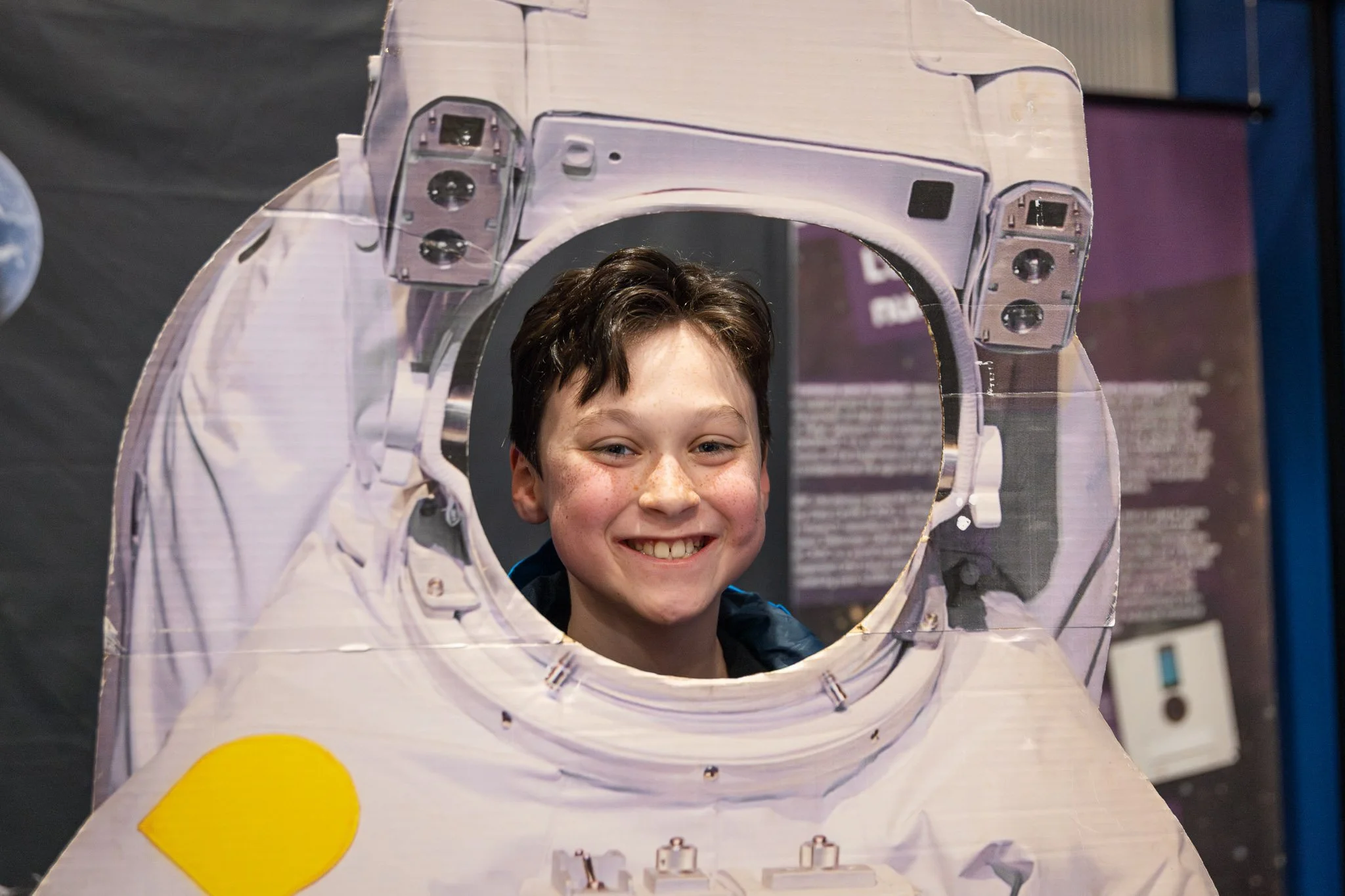 A young boy with short dark hair smiling through a cutout in a cardboard astronaut suit.