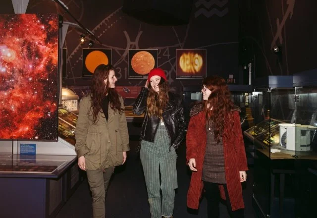 Three young women standing and talking inside a science museum with space and planetary exhibits.