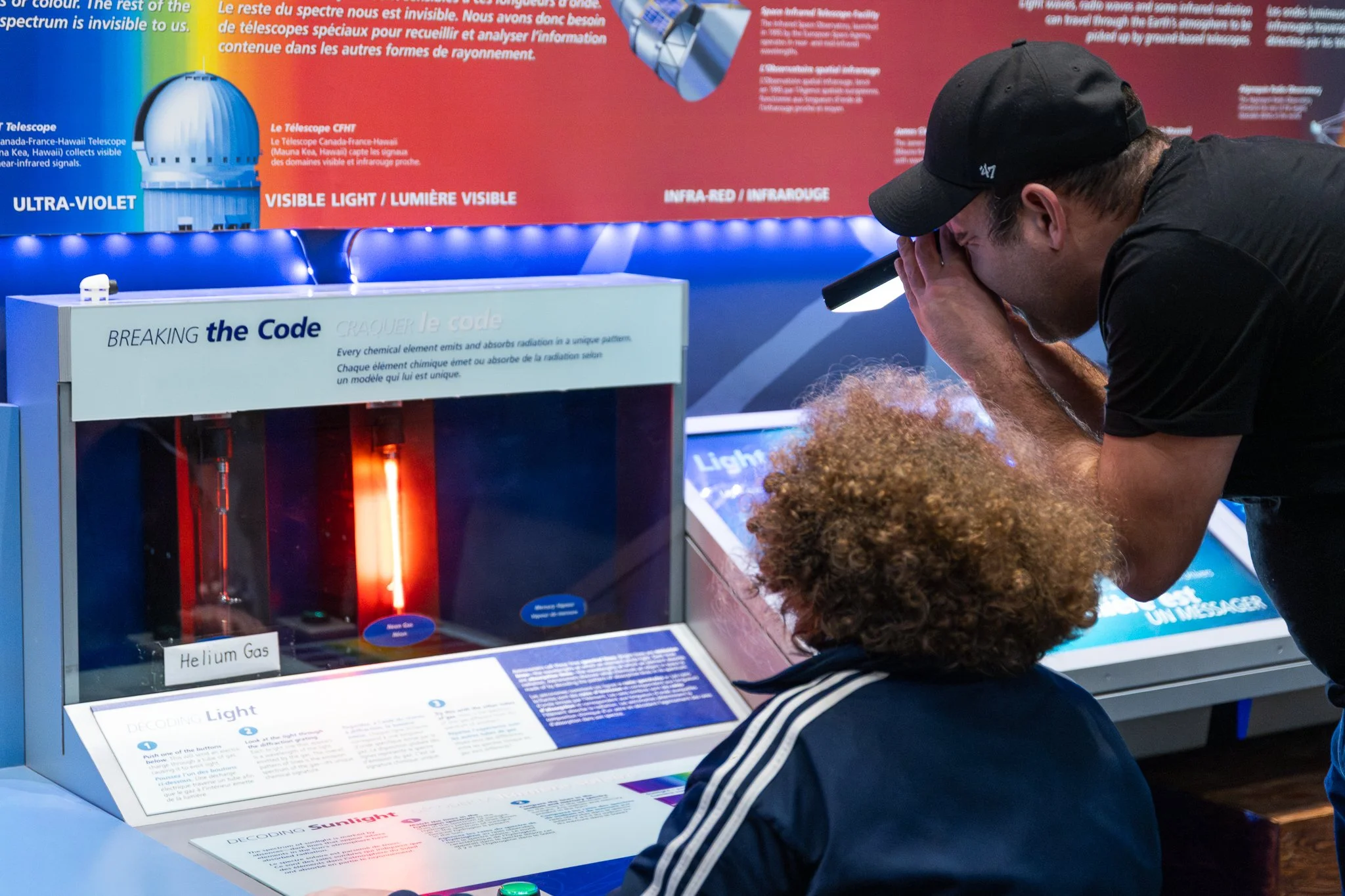 A young boy with curly hair and a dark jacket is looking at an exhibit about the electromagnetic spectrum with a school-aged girl with curly hair, wearing a dark jacket with white stripes on the sleeves. The exhibit features a display titled 'Breakin