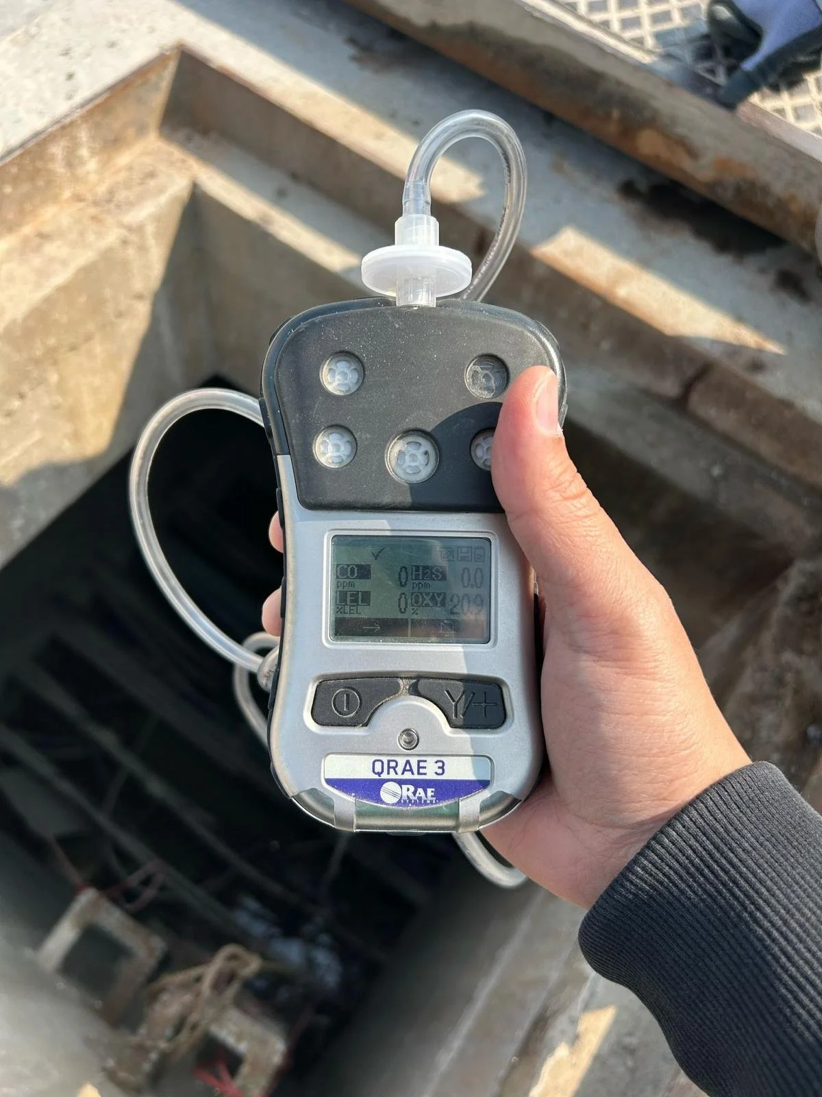 Person holding a radiation detector with a black and silver body, a small screen, and a tube sensor on top, over a construction site.