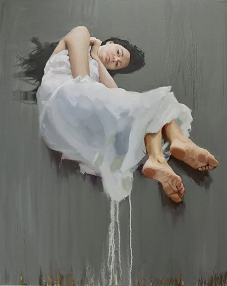 A woman in a white dress lying on a gray floor, with her eyes closed and feet slightly elevated. White paint appears to be pouring from her dress onto the floor.